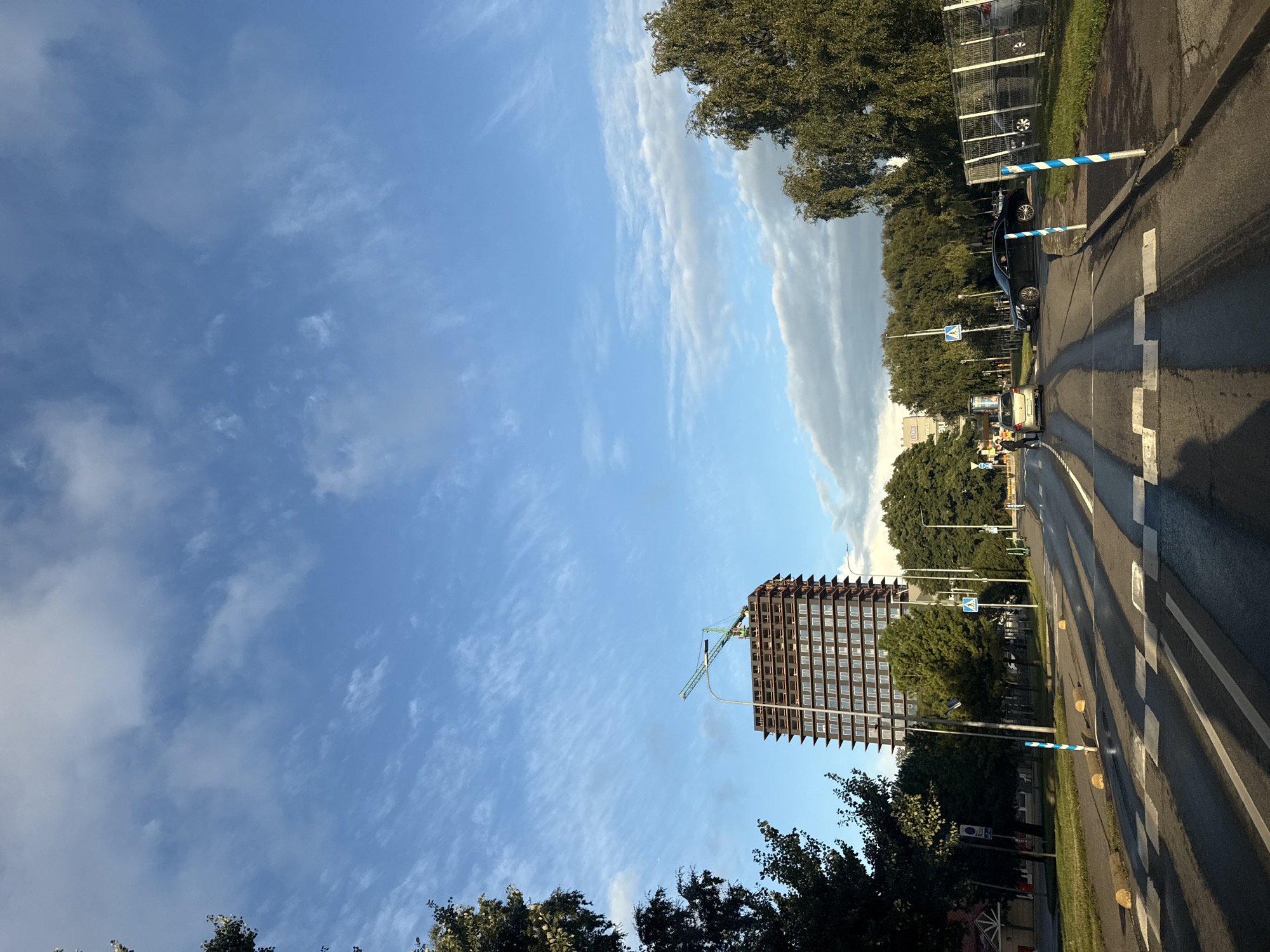 The image captures a street scene in Tallinn, Estonia, under a partly cloudy blue sky. In the background, a tall, unfinished building with a construction crane at its apex is visible. The building is dark brown and has multiple floors with windows. Trees line the street, with some on the left and a denser cluster on the right, partially obscuring a fence.
In the foreground, a wet asphalt road features a pedestrian crossing with white stripes. Cars are present on the road, including a black sedan and a smaller silver car. Two people are standing near the silver car on the left side of the road. The street is marked with lanes and has streetlights. Blue and white striped poles are positioned along the roadside. The overall lighting suggests it is daytime, likely late afternoon due to the long shadows.