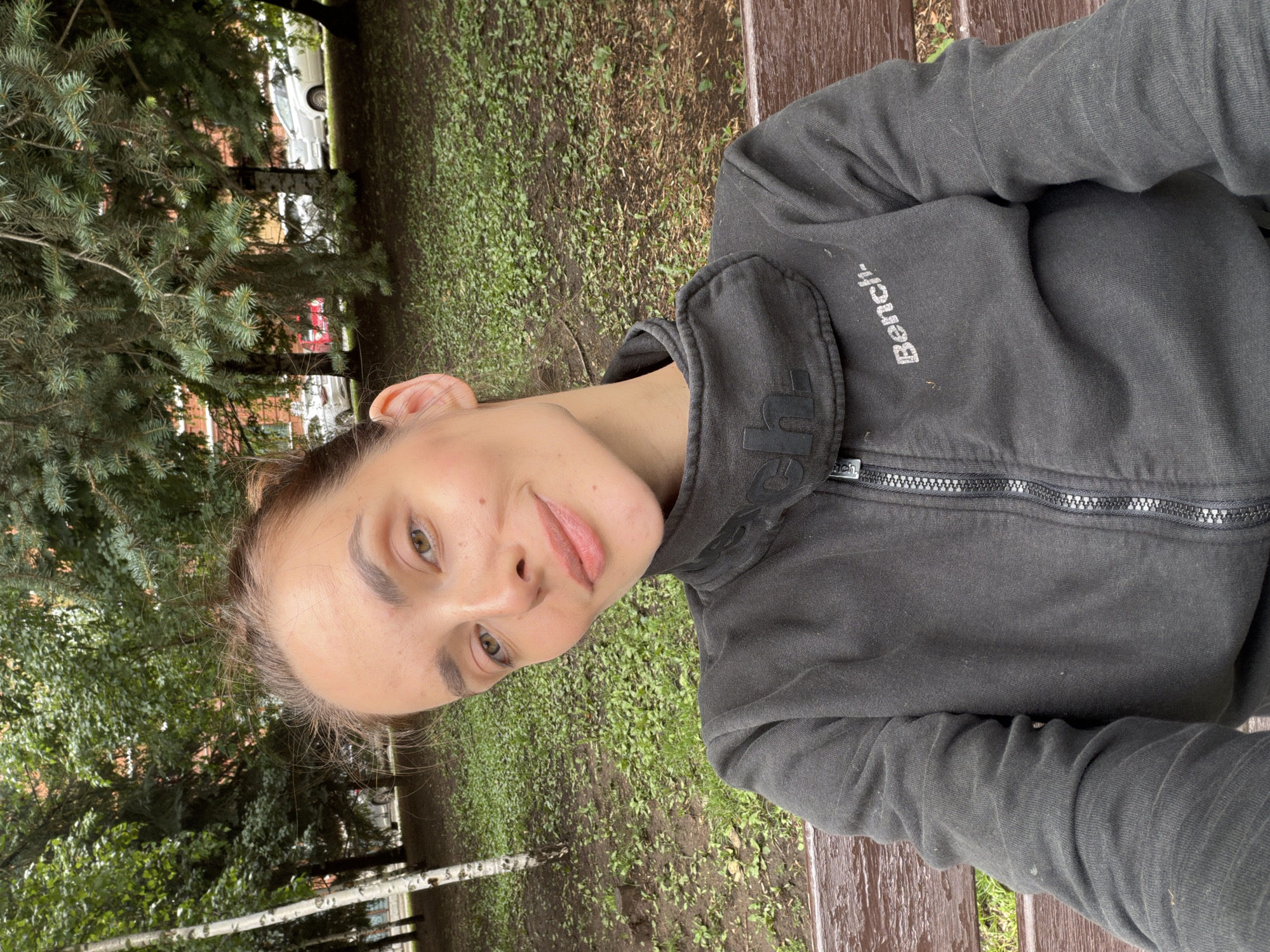 A young woman with light brown hair pulled back and light brown eyes is seated on a wooden park bench, looking into the camera. She is wearing a dark grey zip-up hoodie with a white "Bench" logo on the right side of her chest. The background shows a park setting with green grass, scattered leaves, and trees. In the background, cars are visible parked on a street next to the park, and buildings can be seen through the trees. The image appears to be taken in Ufa, Russia, given the context of the location provided.