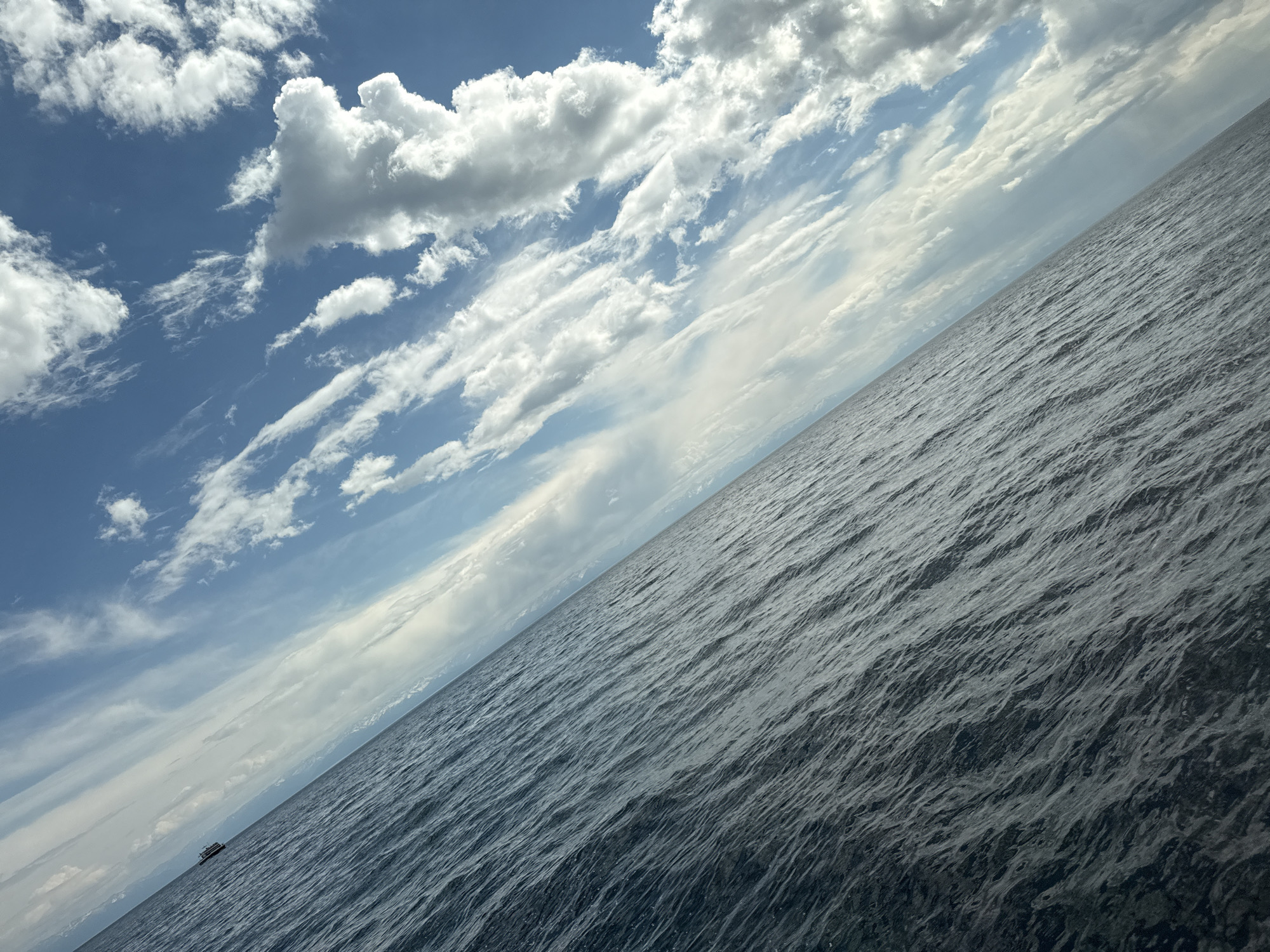 The image shows a body of water meeting a sky with cumulus clouds. The water appears dark and choppy with ripples. The sky has blue patches interspersed with white clouds, ranging from puffy to wispy. A small boat is visible near the bottom left corner of the image. The image is tilted, giving the impression of a diagonal line separating the water and the sky. It is possible to see the location by combining the content in the image with contextual information. Listvyanka, Russia is on Lake Baikal. This scene is likely on Lake Baikal, near Listvyanka.