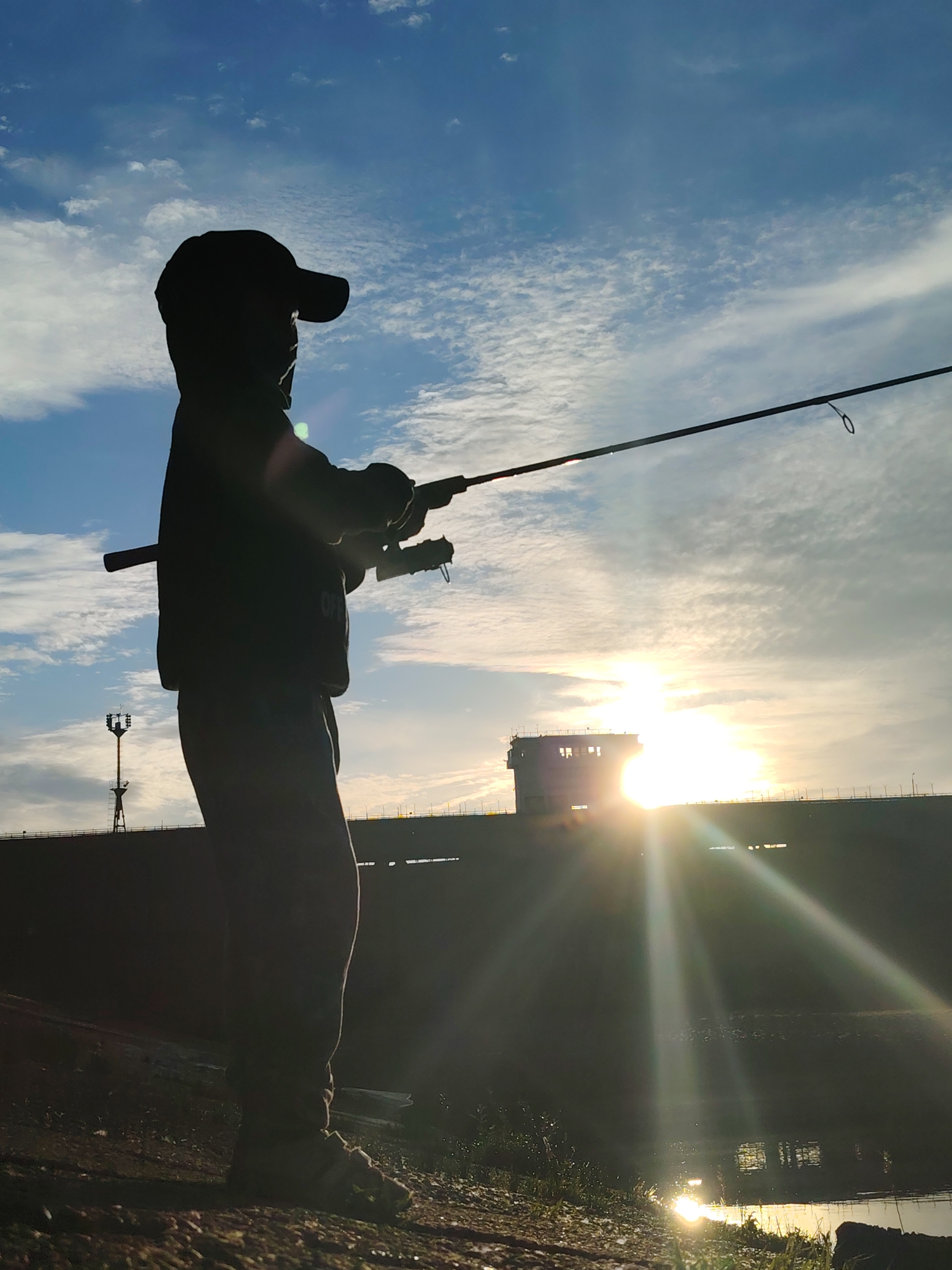 A silhouette of a child wearing a baseball cap and holding a fishing rod stands on the bank of a body of water, facing the sunrise. The sun's rays are visible, creating a lens flare effect. In the background, a concrete structure, possibly a dam or industrial building, is visible. The sky is blue with scattered white clouds. The scene appears to be in Chaikovsky, Russia, given the context.