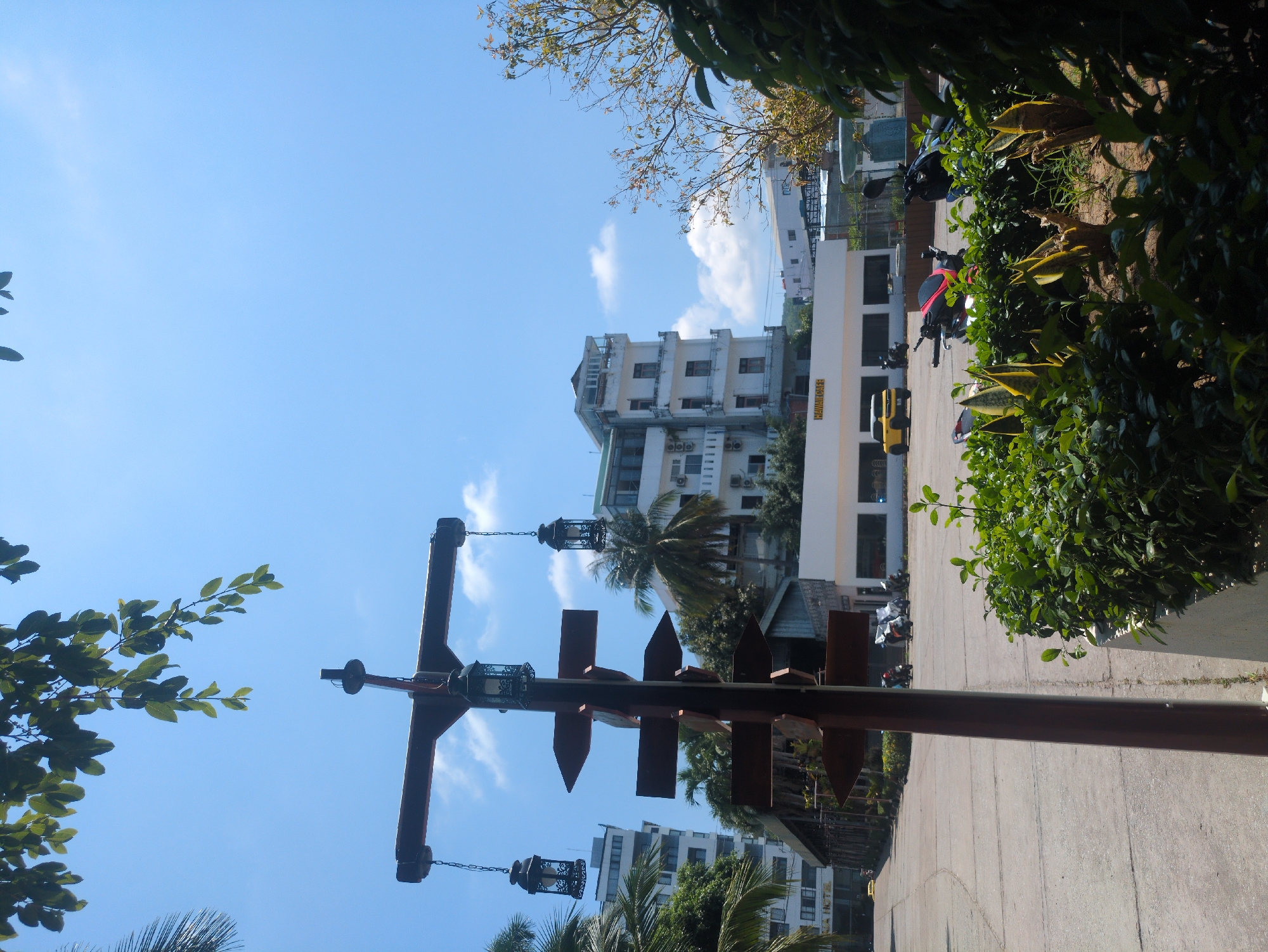 A vertical photograph reveals a street scene in Phú Quốc, Vietnam, showing a combination of architectural elements, vegetation, and street fixtures under a clear sky.
The frame is centered on a tall, ornate, wrought-iron street lamp post that extends upwards and is adorned with hanging lanterns. The post sits on a concrete walkway adjacent to a road.
Behind the lamppost is a multi-story building, characterized by a white facade with evenly spaced windows and visible air conditioning units. To the side of this building, there's a lower, elongated structure that bears the words "HAWAI HOUSE" in yellow letters.
Several parked scooters and a yellow vehicle are visible, suggesting an active street environment. On the right side of the photograph, lush greenery lines the edge of the walkway, partially obscuring the view. The sky is blue with scattered white clouds. Leaves from nearby trees frame portions of the image, providing a natural border.