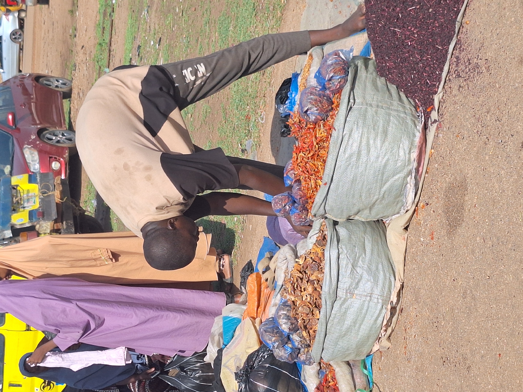 In Kakuri Gwari, Nigeria, a man bends over a roadside market stall. The stall, situated on a paved surface, displays a variety of goods, including dried hibiscus flowers, peppers, and other dried goods packaged in plastic bags and large green bags. The man wears a two-toned tan and black shirt with lettering visible on the sleeve, and black shorts. Two people in traditional gowns stand near the stall. A yellow three-wheeled vehicle and cars are in the background.