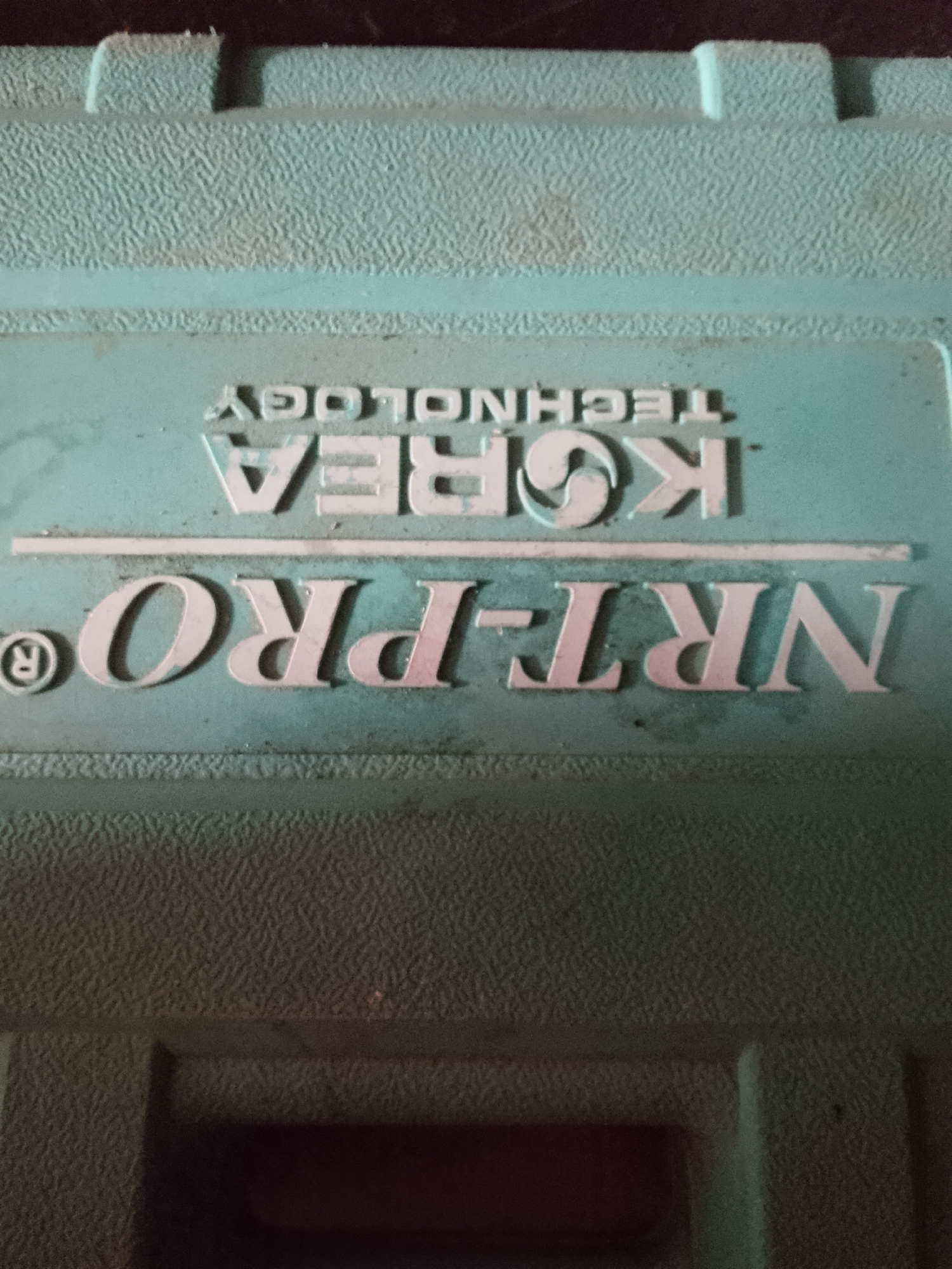 The image shows a close-up, overhead view of a green, textured plastic surface, likely a tool case or equipment casing. Embossed on the surface are two lines of text. The top line reads "KOREA TECHNOLOGY" in all capital letters, with "TECHNOLOGY" appearing upside down. The second line, larger and centered, reads "NRT-PRO®" with the "R" in a circle to the left of "NRT-PRO". The plastic has a slightly worn appearance with some smudges and dust. The background is dark and out of focus, hinting at the surrounding environment. There is no discernible location in the city of Kasiman, Indonesia.