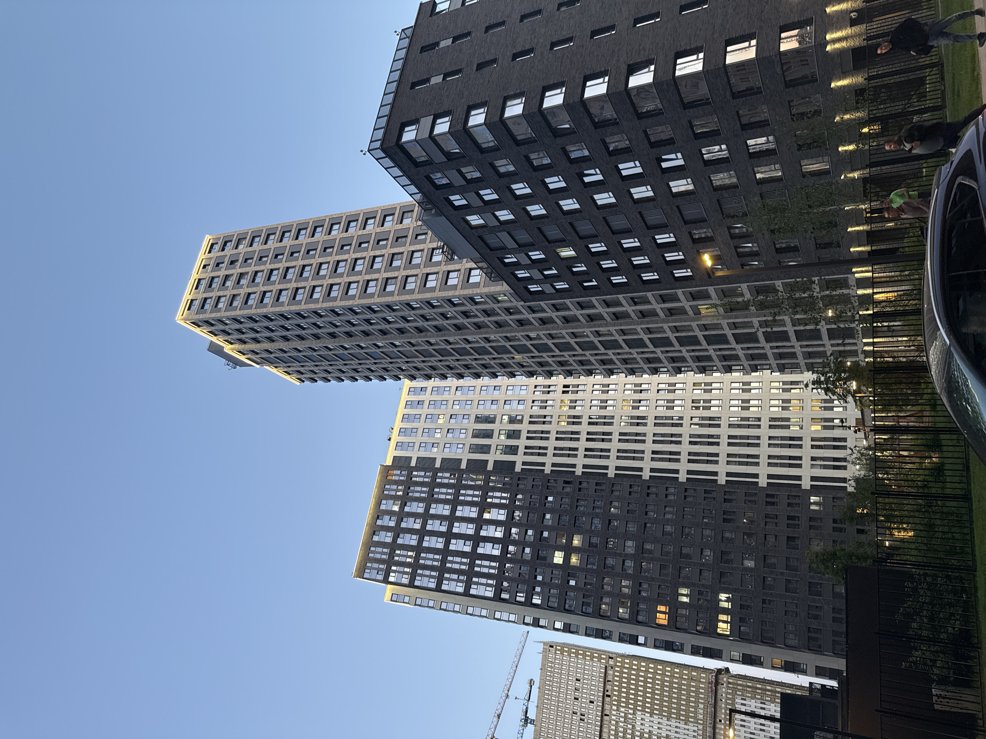 From a low-angle perspective, the image captures several high-rise buildings against a clear blue sky, suggestive of Moscow, Russia. The architecture is modern, with structures featuring grid-like facades and numerous windows. A construction crane is partially visible near the bottom of the frame. On the right side, a fence and partial views of people and a vehicle are visible, adding a human element to the urban scene.