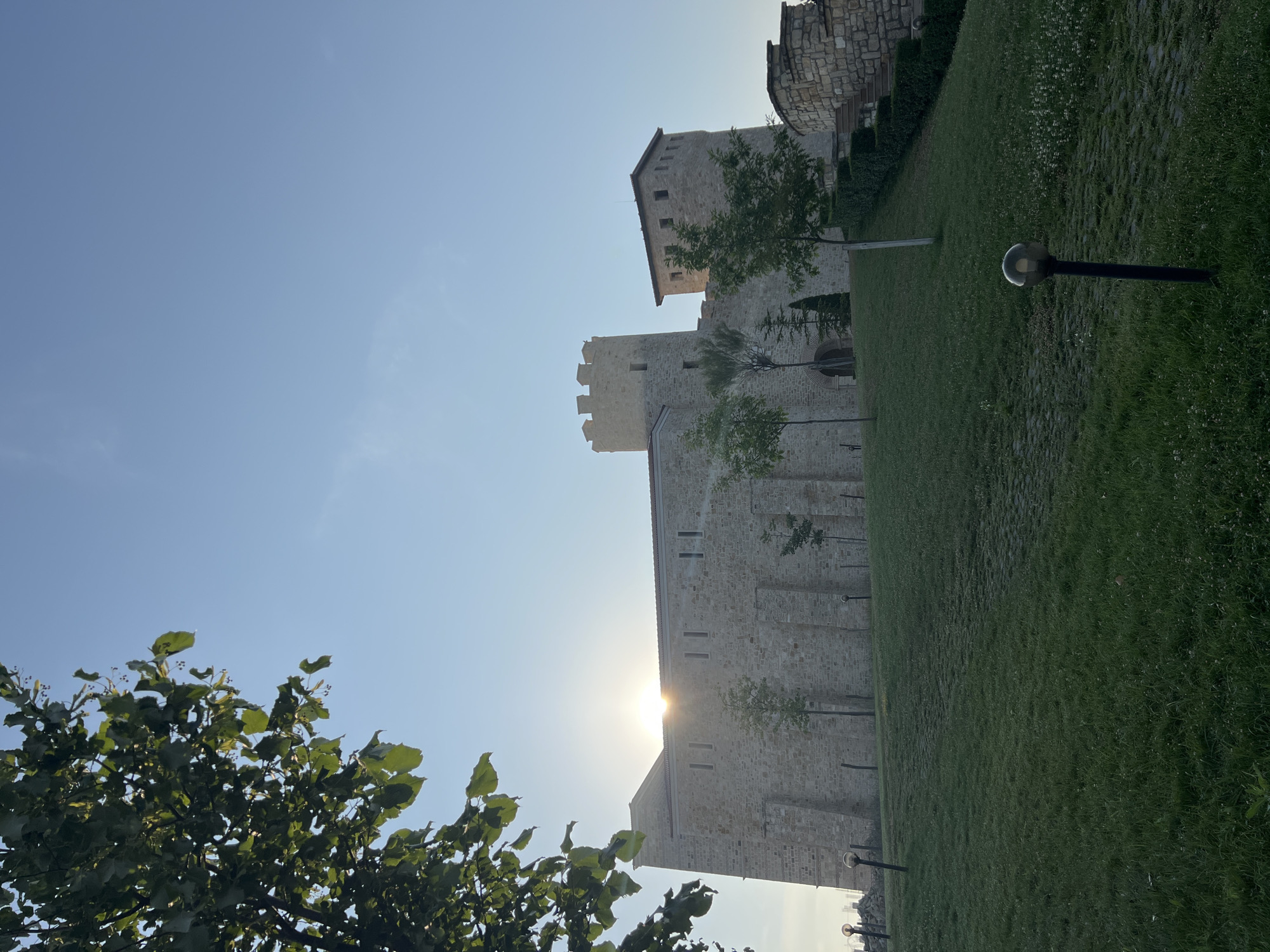 This is a low-angle, full shot image of a stone castle with a clear blue sky. The sun is shining brightly, peeking from behind the castle's roofline. In the foreground, a grassy hill slopes upwards towards the castle, with a stone pathway visible on the grass. Several trees and bushes are scattered on the grounds. Two lampposts with globe-shaped lights are planted on the hill. To the left, a tree branch with green leaves hangs into the frame. The castle appears to be a historical structure with towers and crenellated battlements. The overall scene is illuminated by the sun, creating a warm and slightly hazy atmosphere. The location is identified as the fortress in Neofit Rilski, Bulgaria.