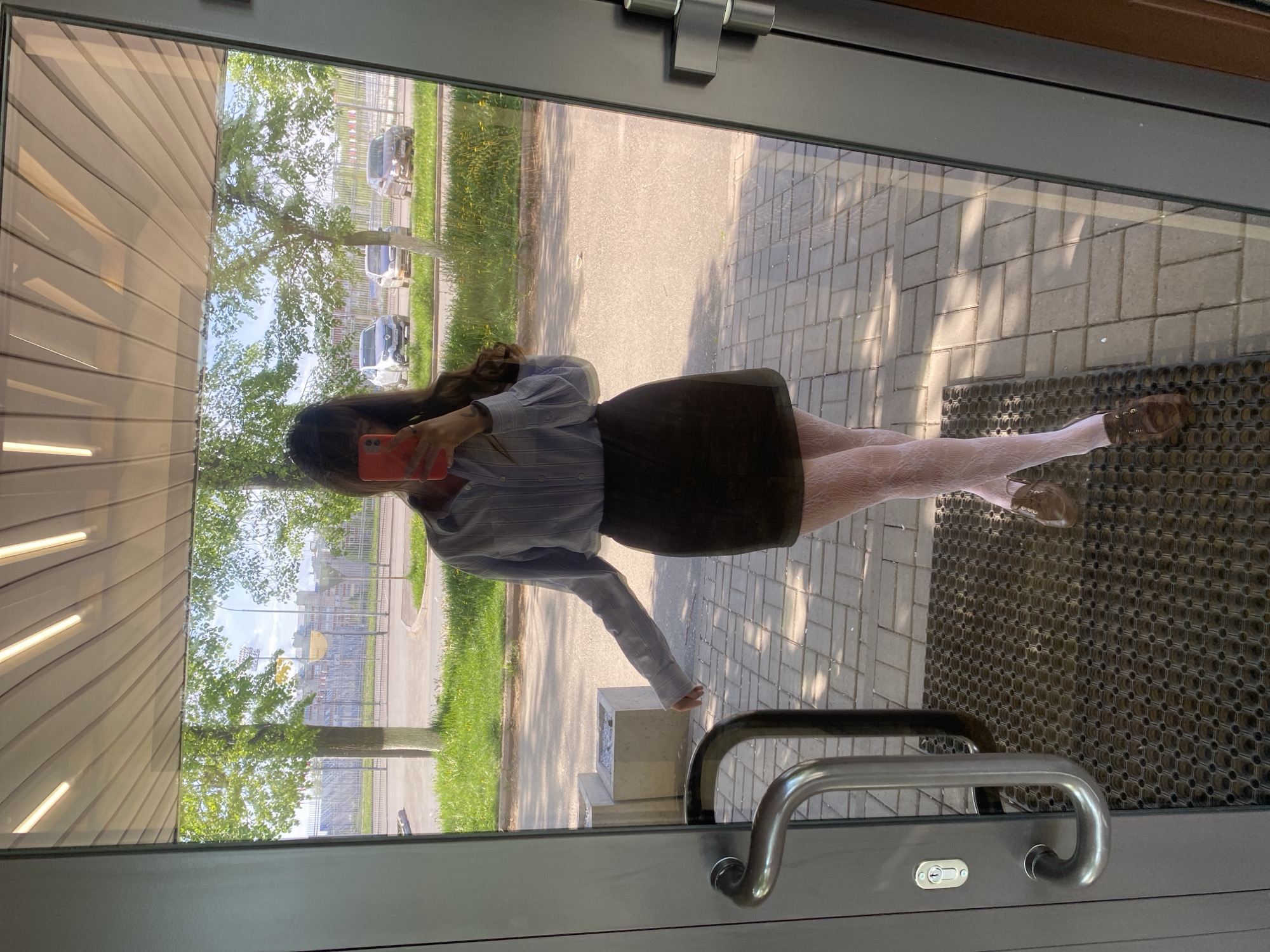 A woman is taking a selfie in a doorway in Votkinsk, Russia. She is wearing a striped shirt, a dark skirt, and patterned tights. Outside the doorway, there is a paved area, grass, trees, and parked cars. The reflection in the doorway shows a building with horizontal lines. A door handle is visible in the lower right corner.