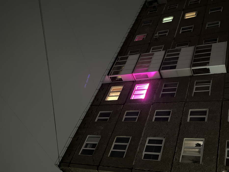 This image depicts a multi-story residential building at night in Neftekamsk, Russia, under an overcast, dark gray sky. The building features a dark, textured facade, likely concrete or stucco, with numerous rectangular windows, most framed in white. A prominent, intense magenta light emanates from a window on the third floor (from the bottom), located to the immediate left of a stack of vertically aligned, enclosed white balconies. This magenta light casts a distinct glow onto the adjacent section of the enclosed balcony. Other windows display varying interior illumination, including warm yellow and cooler white lights, while many remain dark. Thin, dark utility lines are visible stretched across the sky above the building. A faint, segmented purple streak is present in the sky, positioned horizontally above the building. A dark, leafy plant is partially visible in the bottom-left window on the first floor. No individuals or specific actions are discernible.