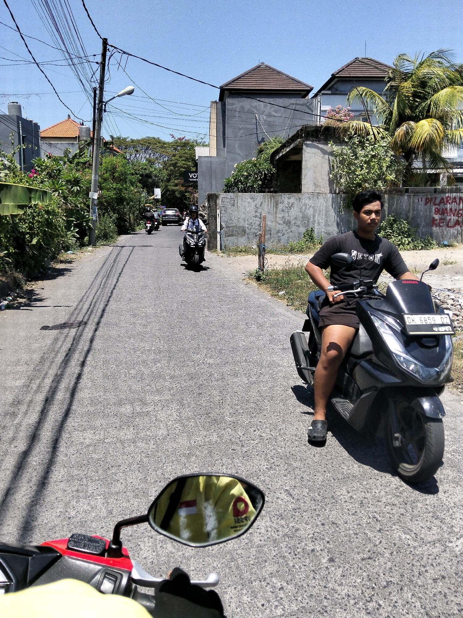 The image captures a street scene in Denpasar, Indonesia, on a sunny day. A young man is seated on a black scooter, looking towards the viewer. He is wearing a black t-shirt with white lettering and dark shorts, with black crocs on his feet. The scooter has a license plate that reads "DK 6859 DZ". Behind him, other scooters and a car are visible on the road, suggesting moderate traffic. The street is lined with buildings, some with traditional tiled roofs, and lush green vegetation. Power lines crisscross overhead, a common sight in urban Indonesian landscapes. In the foreground, the reflection in the scooter's mirror shows a person in a yellow shirt with a red and white Indonesian flag visible. The overall atmosphere is one of everyday life in a bustling Indonesian city.