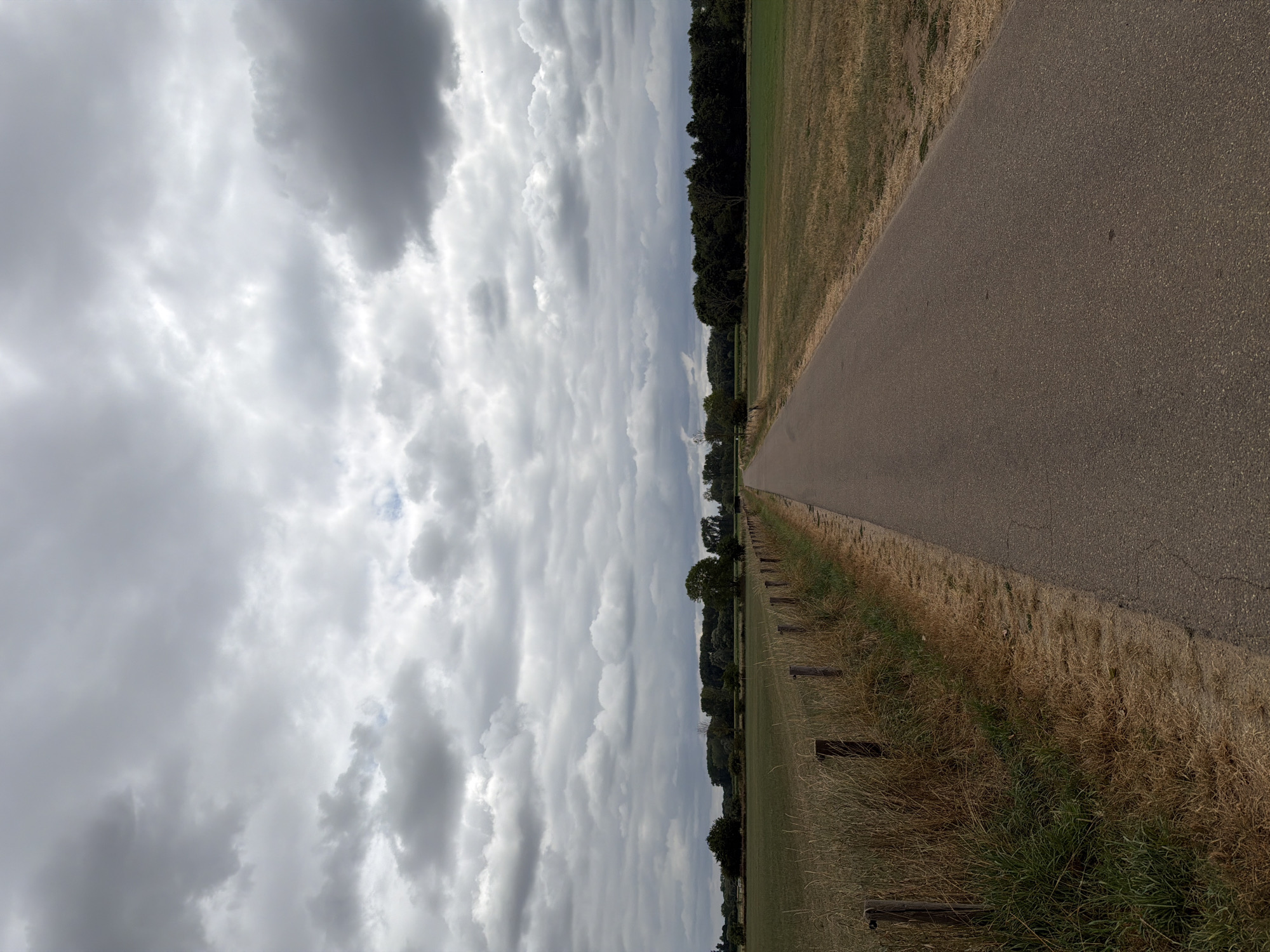 The image depicts a paved pathway in Melick, Netherlands, running through a rural landscape. The path is flanked on the left by a dry, grassy verge with a rustic wooden fence and beyond that, a field of tall, dry grass. To the right of the path, there is a cultivated field with green grass. In the background, a line of trees forms a dark green border against the horizon. The sky is overcast with heavy, grey clouds. There are no people or animals visible in the frame. The scene appears quiet and undisturbed.