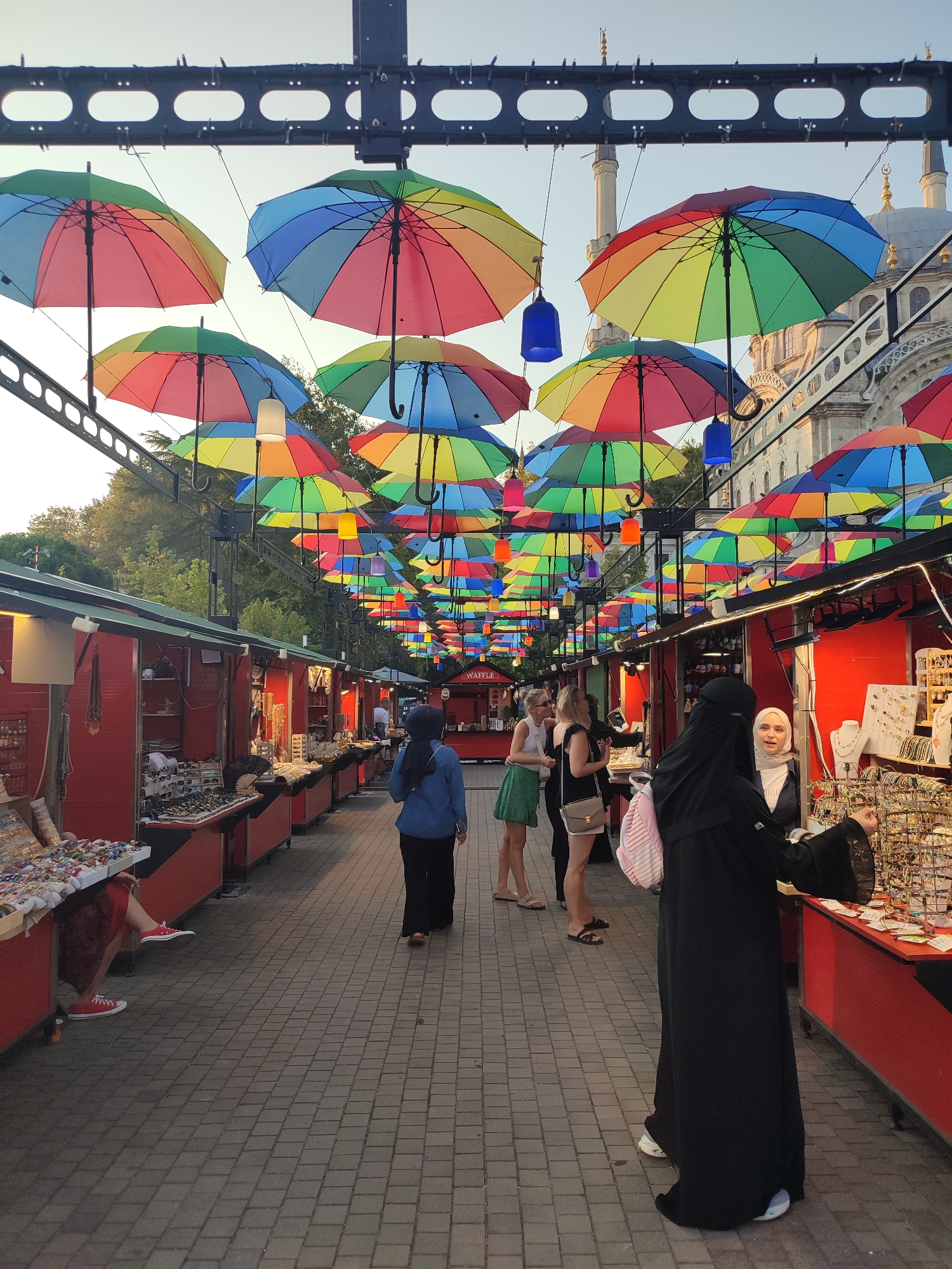 The image captures a vibrant market scene in Beyoğlu, Turkey, beneath a canopy of colorful, rainbow-patterned umbrellas suspended from a metal structure. The market features rows of red stalls displaying an assortment of goods, including jewelry, trinkets, and other retail items. In the foreground, several individuals are browsing the merchandise. On the left, a person in a red skirt and red sneakers is seated, partially obscured. To the right, two women in casual attire are looking at a stall. Further down the market lane, a person wearing a dark headscarf and a blue jacket walks away from the camera. Towards the right side of the image, two women, one in a black abaya and headscarf, and another in a white hijab, are interacting at a stall laden with jewelry. The background reveals the architecture of a building with domes and minarets, likely a mosque, characteristic of Istanbul. The overall ambiance suggests a lively daytime market setting with a decorative overhead display.