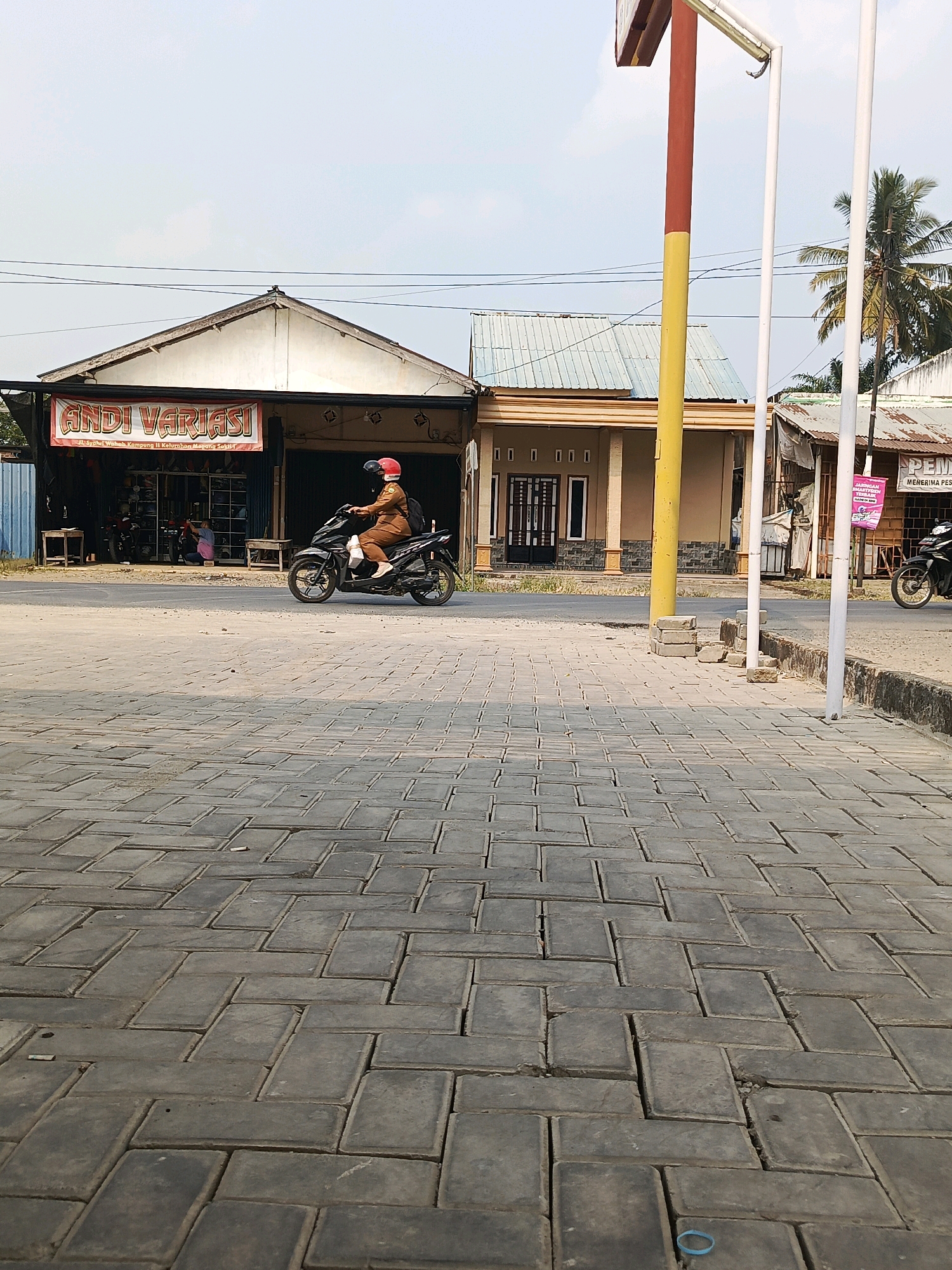 The image captures a street scene in the city of Megang Sakti I, Indonesia. In the foreground, a paved walkway made of interlocking grey rectangular bricks stretches towards the viewer, with some bricks slightly uneven. A blue rubber band is visible on the walkway in the lower right corner.
The midground features a road with a person riding a black motorcycle from left to right. The rider, wearing a brown outfit and a red and blue helmet, is positioned on the motorcycle, which has its headlights on. The motorcycle is passing in front of several buildings.
To the left of the road, there is a shop with a prominent red and white banner above its entrance that reads "ANDI VARIASI" in bold lettering. Below the banner, smaller text indicates the address: "Jl. Syaiful Wahab, Kampung II, Kelurahan Megang Sakti". The shop's interior appears to display various items, and two individuals can be seen inside. Two wooden stools are placed outside the shop on the left and right.
To the right of the "ANDI VARIASI" shop, another building with a light brown facade and a metal roof is visible. It has several rectangular openings and a dark-colored door. Further to the right, a taller building with a corrugated metal roof and a pink banner is situated.
A prominent yellow and red pole with a white top stands vertically in the midground, to the right of the "ANDI VARIASI" shop, and a white pole is next to it. The background shows a clear sky with some faint clouds, and a palm tree is visible to the far right. Power lines are strung across the sky above the buildings. The overall environment suggests a typical urban or semi-urban setting in Indonesia, with a focus on local businesses and daily activities.