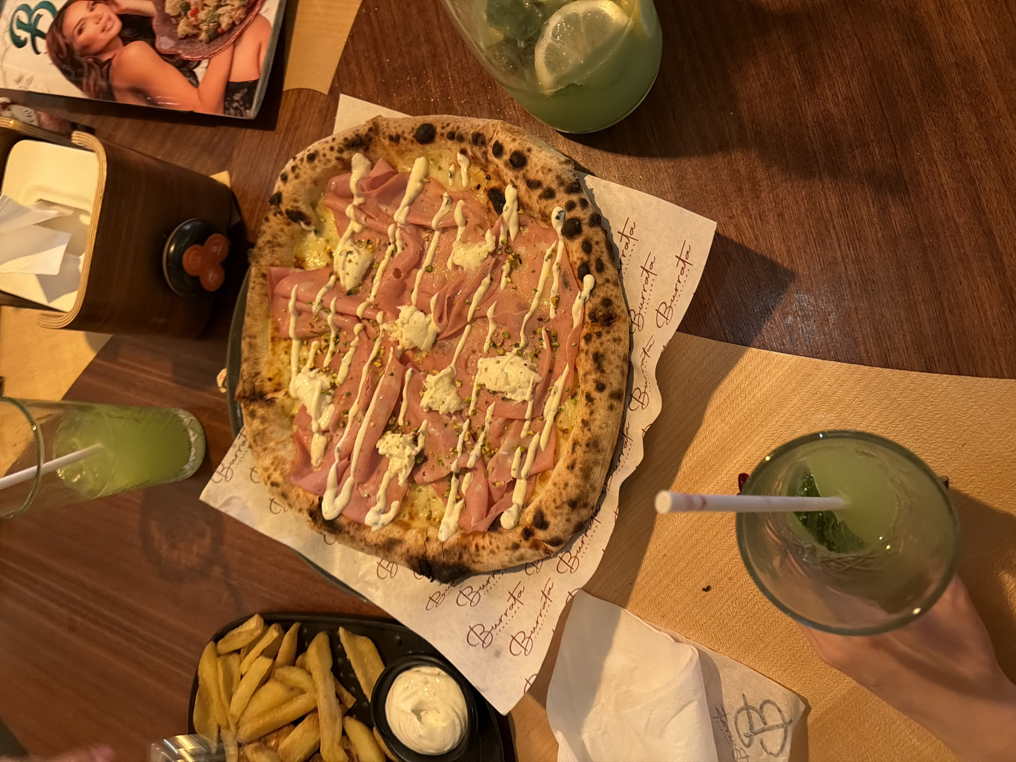 The image captures a top-down view of a dining table at the Burrata Italiana restaurant in Sofia, Bulgaria. Central to the scene is a large, Neapolitan-style pizza featuring a blistered crust, generously topped with thinly sliced mortadella, several dollops of burrata cheese, and a light sprinkling of chopped pistachios. The pizza rests on white parchment paper bearing the "Burrata Italiana" logo.
To the immediate left of the pizza, a black rectangular serving dish holds a portion of thick-cut French fries. Adjacent to the fries is a small, black ramekin containing a white, creamy sauce or dip.
Several beverages are visible:
1. In the upper left, a tall, clear glass with a white straw contains a translucent green liquid.
2. To the right of the pizza, a clear glass bottle filled with a similar green liquid is seen, containing lemon slices and mint leaves.
3. In the bottom left, a hand with painted nails holds another tall, clear glass with a white straw, also containing a translucent green liquid.
Further objects on the dark wooden table include a rectangular wooden napkin dispenser filled with white paper napkins, positioned in the upper right quadrant. Partially visible next to the napkin dispenser is a magazine or menu with a woman on the cover, holding a bowl of food, and the letter "B" prominently displayed at the top. The overall environment is consistent with an indoor restaurant setting. No specific actions beyond the presumed act of dining are depicted, though the presence of multiple food and drink items suggests a shared meal.
