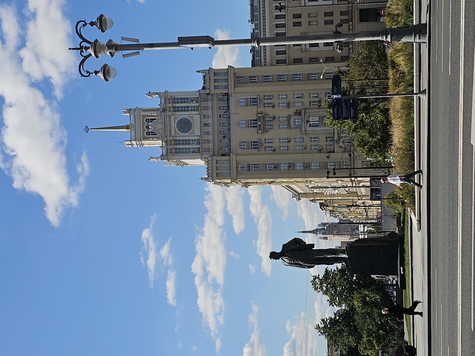The image depicts a street scene in Moscow, Russia, featuring a prominent statue in the foreground and a distinctive Stalinist skyscraper in the background. The statue, a dark silhouette of a man in a suit, stands on a large, dark pedestal. To the right of the statue, a tall, ornate lamppost with three lights is visible. Behind these elements, a large, cream-colored Stalinist skyscraper dominates the skyline. It features numerous windows, decorative architectural details, a clock face, and a spire topped with a star. Other buildings, some with similar architectural styles, line the street. The sky is blue with scattered white clouds. Two individuals are walking on the sidewalk in front of the statue and buildings. One person, dressed in dark clothing, is walking from left to right, and another, wearing a white shirt and dark pants, is walking from right to left. A roadway with lane markings is in the foreground. The overall environment appears to be urban, with trees and landscaping present around the statue and along the sidewalk.