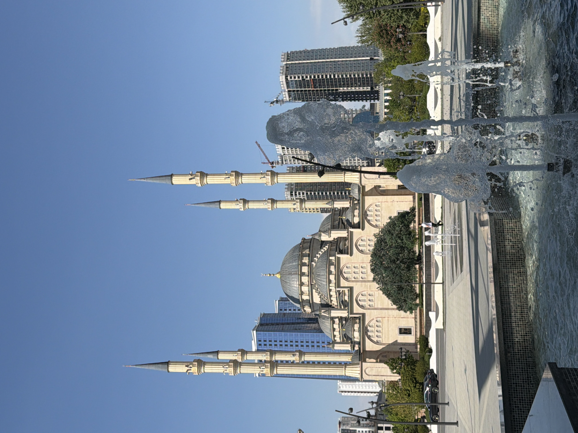 The image depicts the Akhmad Kadyrov Mosque in Grozny, Russia, observed from an eye-level perspective beside a fountain. The mosque, constructed with light beige stone, features a central dome and four minarets with pointed spires. Water jets from the fountain are visible in the foreground, partially obscuring the mosque. In the background, modern high-rise buildings, some under construction, suggest the city's urban environment. The sky is a clear blue.