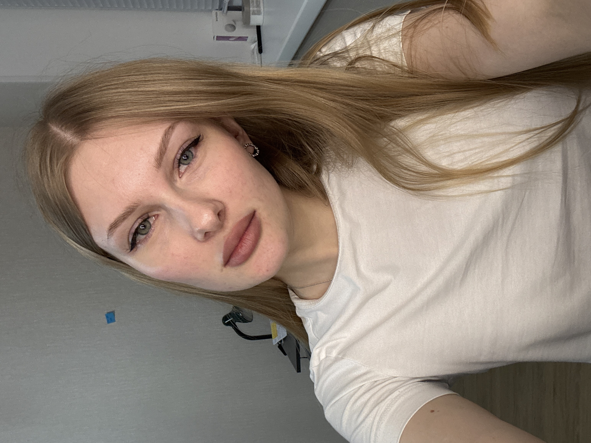 A young Caucasian woman with long, straight, light brown hair and light green eyes is featured in a close-up, rotated selfie. She looks directly at the camera with a neutral, posed expression. She is wearing a plain white short-sleeved t-shirt, a small circular silver-colored earring in her right ear, and a thin, light-colored chain necklace. Her lips are full and tinted with a light pink/nude color, and she has subtle eyeliner. The background suggests an indoor setting, consisting of a plain light grey wall, where a small blue square sticker is visible, and a white ceiling with some indistinct white and dark objects, possibly light fixtures or machinery. A small reddish-brown mark is present on her bare arm. No text is visible in the image.