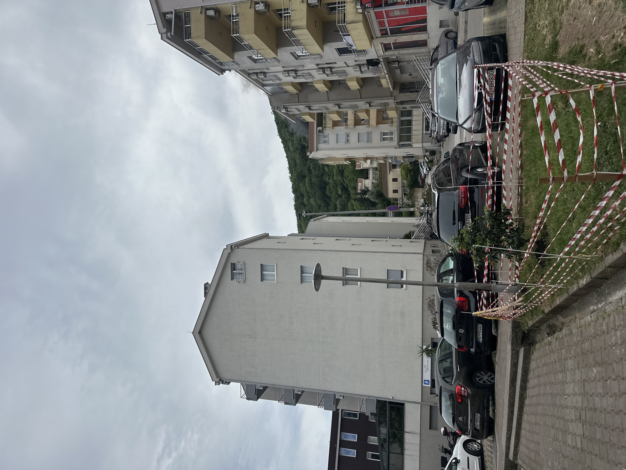 The image shows a street scene in Budva, Montenegro. Several multistory buildings are visible, some with balconies. The sky is overcast. Parked cars line the street along a sidewalk paved with interlocking stones. Red and white barrier tape surrounds a grassy area adjacent to the sidewalk. The hills are forested in the background.