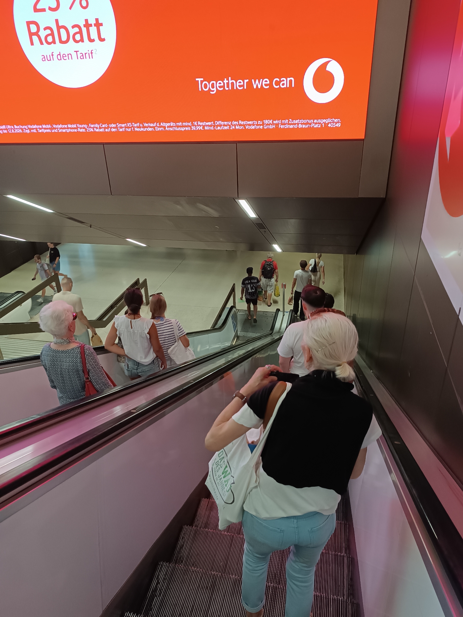 The image captures a dynamic scene on an escalator in a modern public space, likely a subway station or a shopping mall in Munich, Germany. Dominating the top half of the frame is a large, orange digital advertisement from Vodafone, featuring a prominent "25% Rabatt auf den Tarif" (25% discount on the tariff) and the slogan "Together we can". Below the advertisement, a group of people are ascending on an escalator. In the foreground, a woman with white hair, wearing a patterned blouse and a red handbag, is looking towards her left. Behind her, other individuals are also on the escalator, including a woman in a white top and denim shorts, and another in a striped t-shirt with a white tote bag. Further down the escalator, more people are visible, some descending and some ascending, creating a sense of movement and transit. The environment is characterized by clean, metallic surfaces of the escalator and a muted, contemporary architectural design. The lighting suggests an indoor setting, with overhead lights contributing to the overall brightness. The advertisement, with its distinct Vodafone branding, is a key element indicating the location is likely a commercial area within Munich where such promotions would be displayed.