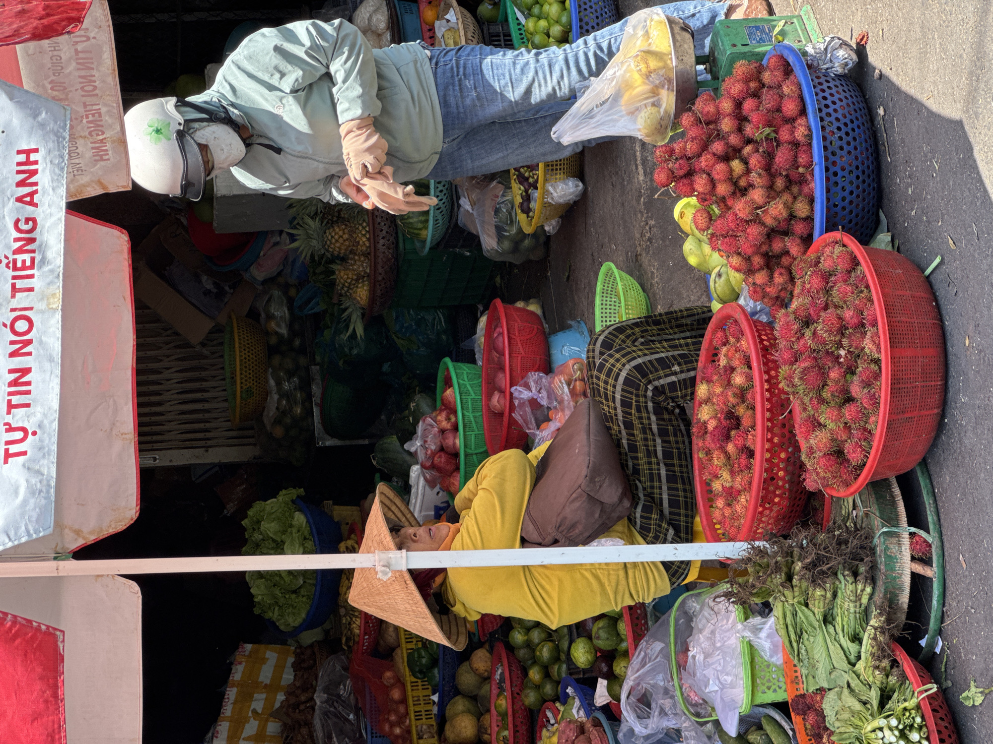 The image depicts an outdoor market scene in Nha Trang, Vietnam. A person wearing a light green jacket, blue jeans, a white helmet with a green flower emblem, and a white face mask is standing, holding a pair of light brown gloves. They are in the process of putting on the gloves. Next to them, a person wearing a yellow long-sleeved shirt and a conical straw hat is sitting, with a brown bag on their lap. They are surrounded by various fruits and vegetables displayed in baskets and crates. In the foreground, two red baskets are filled with rambutan, and a blue basket is also filled with rambutan. To the left, a bundle of leafy green vegetables is visible. In the background, there are more displays of fruits and vegetables, including pineapples, limes, and what appear to be apples. The market stall is covered by a red and white awning with Vietnamese text on it. The ground appears to be asphalt.