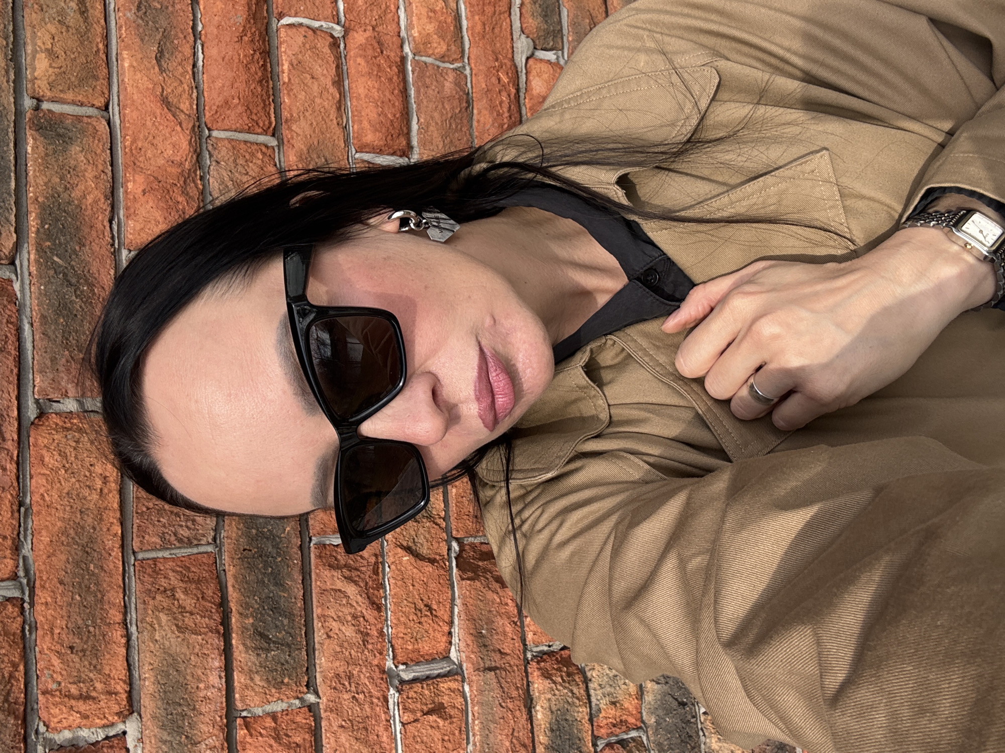 This close-up, diagonal selfie captures an adult individual, likely in Chaikovsky, Russia, posing against a textured red brick wall. The person has long, straight black hair and fair skin, with their head tilted. They are wearing large, rectangular black sunglasses, which obscure their eyes. Their lips are slightly parted, revealing a light pink lip color.
The individual is dressed in a tan or khaki-colored jacket, possibly a utility or workwear style, over a black shirt. Notable accessories include a distinct silver-toned, geometric earring worn on the left ear, and a silver watch with a rectangular light-colored face on their left wrist. A simple silver band ring is visible on the ring finger of their right hand, which is gently placed on the jacket near the chest.
The background consists of a well-defined red brick wall with gray mortar, suggesting an outdoor location or an interior with exposed brickwork. The lighting indicates it is daytime, with clear, possibly bright, light. There is no visible text in the image.