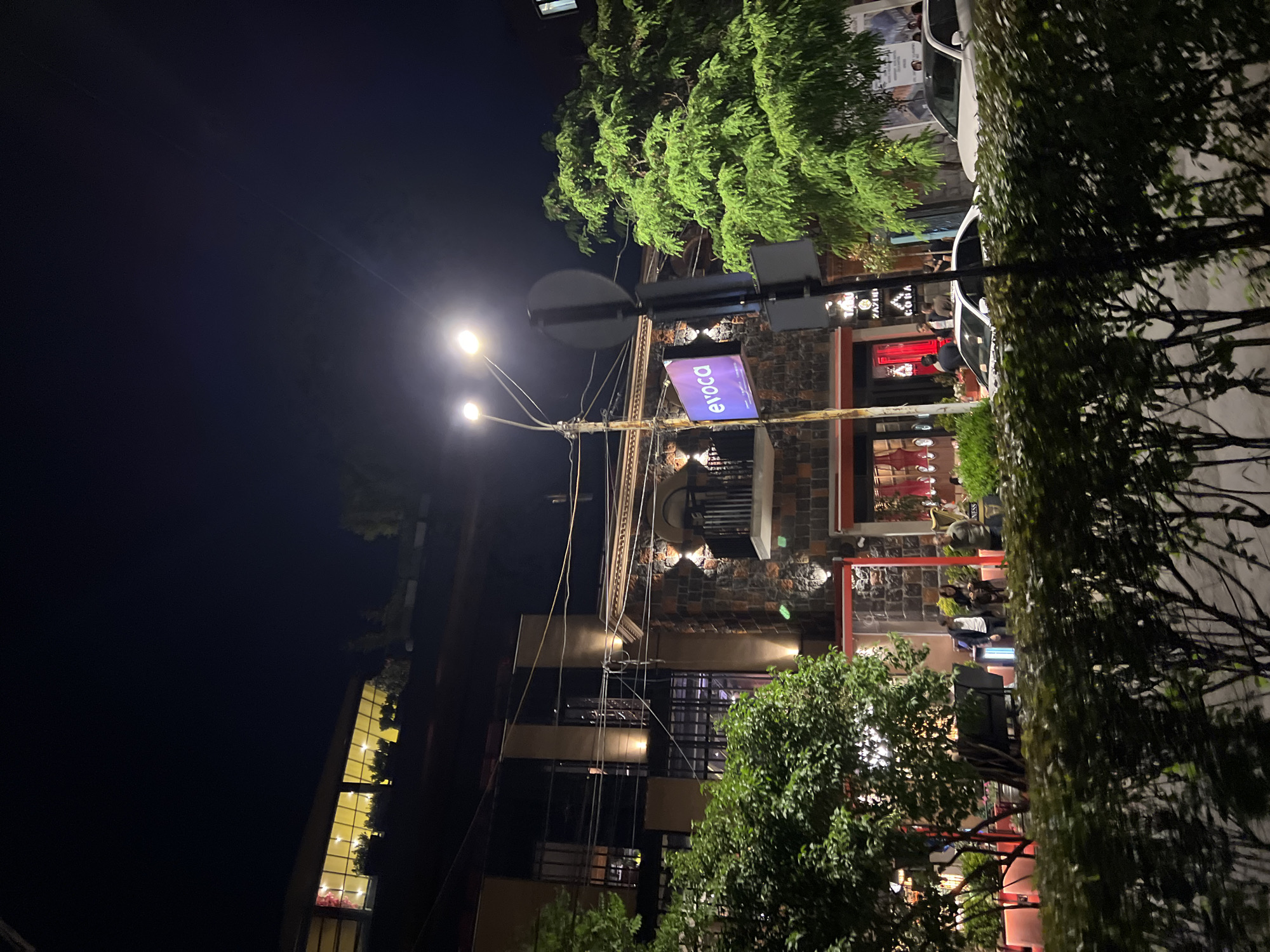Captured at night in Yerevan, Armenia, the scene presents a street view focusing on a building with distinct architectural elements. The building features brick and stone facade, several window levels, and visible lighting both inside and outside. A street lamp illuminates the area, adding to the ambient light. A street sign with the inscription "evoca" is prominently displayed. Trees and other greenery partially obscure some sections of the building. People can be seen in the area near the building entrance or patio. The overall environment suggests an urban setting during nighttime.