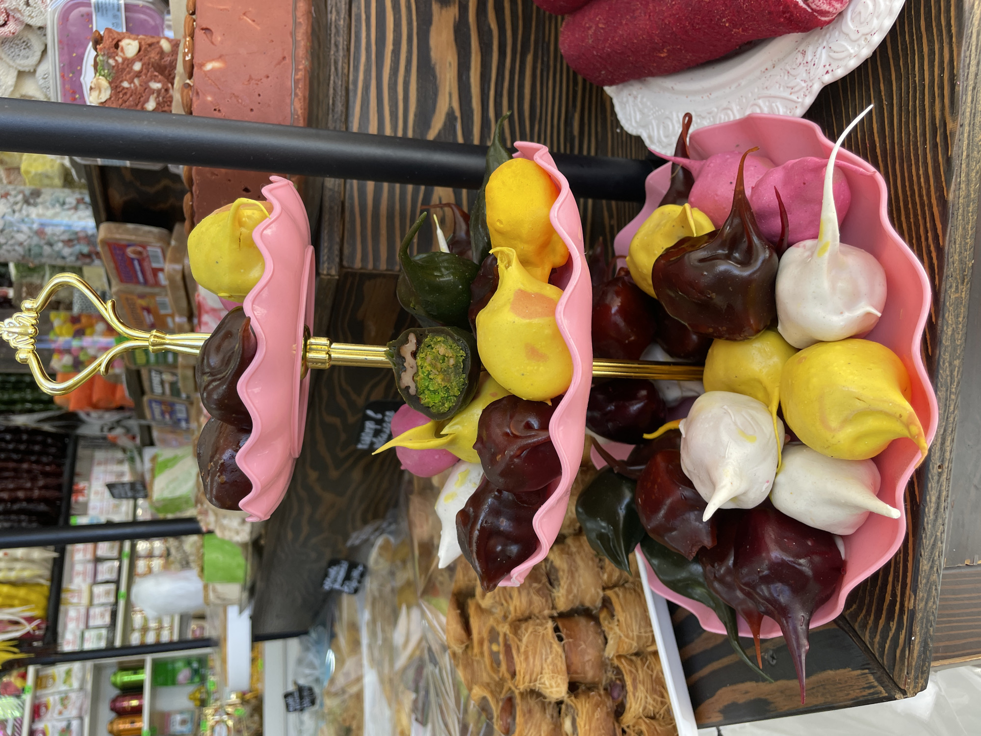 A close-up shot captures a two-tiered golden stand adorned with pink, lotus-shaped bowls. These bowls are filled with an assortment of colorful confections or decorative edibles, primarily in shades of dark red/brown, vibrant yellow, white, pink, and green. Many of these items are pear-shaped or feature distinctive pointed tips.
The stand rests on a dark wooden surface, possibly a counter or display case, against a backdrop of a wooden wall. In the upper left background, a clear container holds a reddish-brown confection, resembling halva or nougat, studded with nuts. To the upper right, a red, cylindrical food item, possibly a type of sausage or pressed meat, is presented on an ornate white plate. Below the stand, a tray filled with intricate pastry, likely kadayif or baklava, made from shredded phyllo dough, is visible. The deeper background reveals blurred shelves stocked with various packaged goods, suggesting the setting is a sweet shop or a market stall. The scene portrays a rich and varied display of specialty food items. No discernible text is clearly legible.