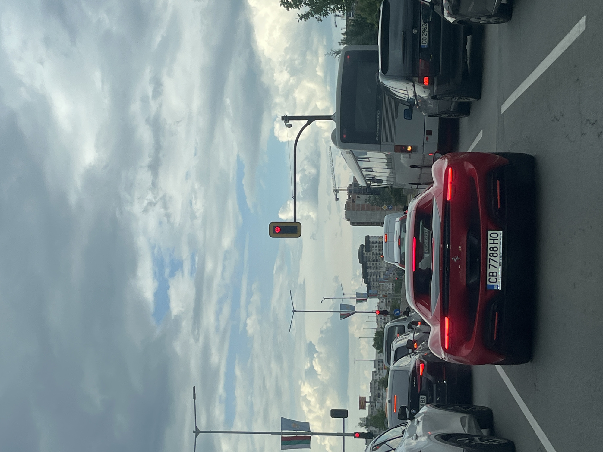 The image shows a traffic jam on a street in Sofia, Bulgaria. The sky is cloudy. A red Ferrari with the license plate CB 7788 HO is prominently featured. Several other vehicles, including a bus with license plate CO 2982, are visible in the traffic. Traffic lights are red. Bulgarian flags are on lampposts along the road. A construction crane is visible in the background.