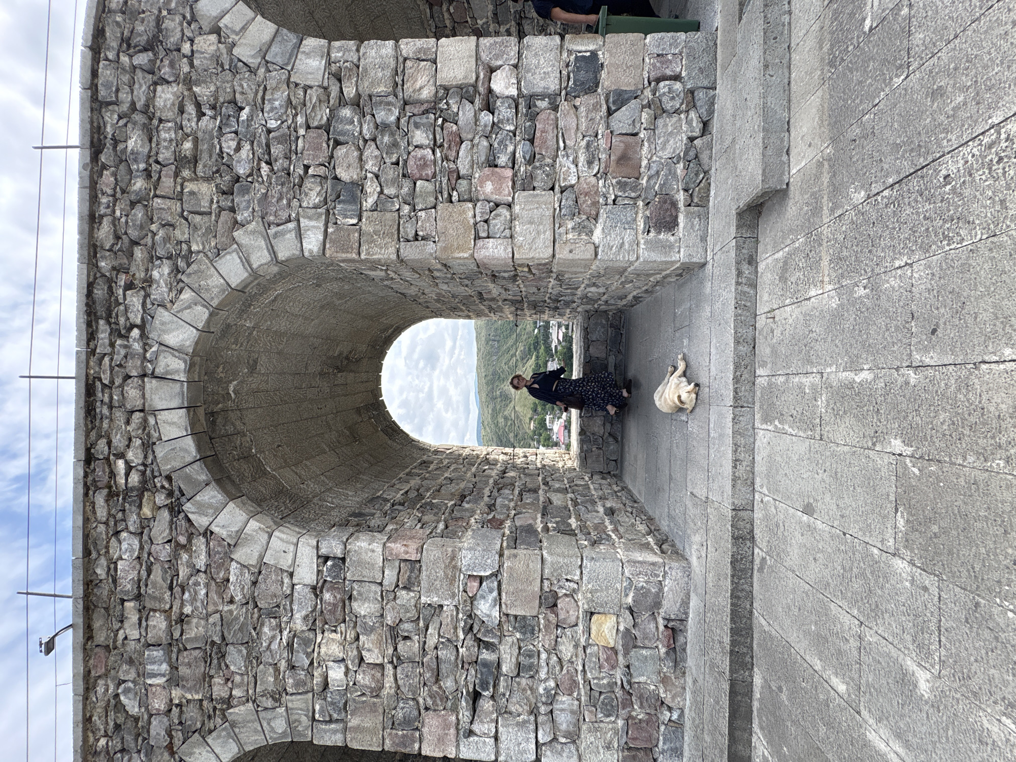 In Akhaltsikhe, Georgia, a person wearing a black dress with a floral pattern and a dark cardigan stands within an arched stone passageway. They are looking towards a distant landscape of rolling green hills. A light-colored dog lies on the stone pavement in front of the person. The passageway is constructed from rough-hewn stone blocks of various sizes and colors, with arched openings framing the view of the outside. The ground in front of the passageway is paved with large, grey stone slabs. The sky above is partly cloudy.