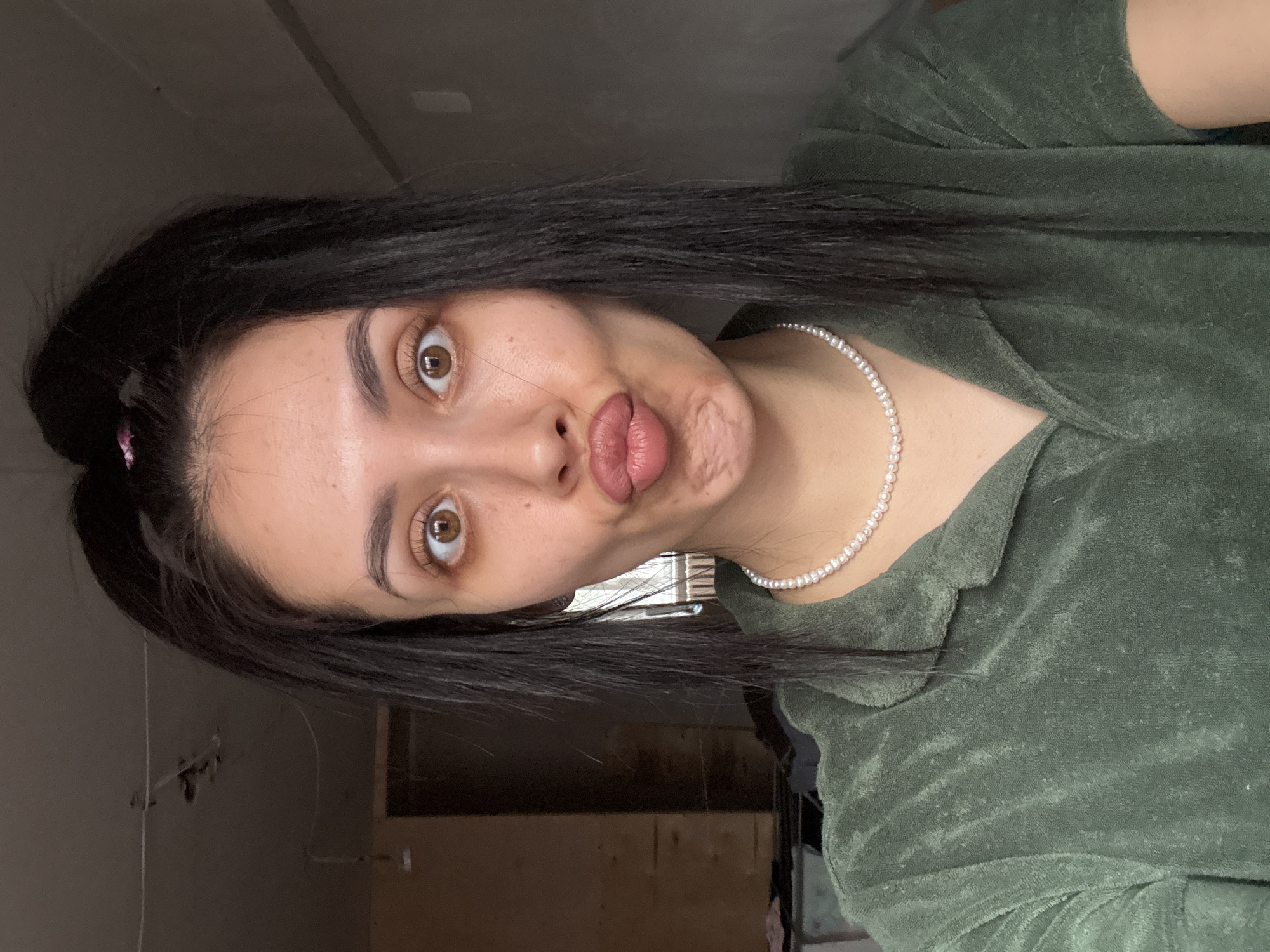 A close-up selfie, taken indoors in Votkinsk, Russia, features a young woman with a playful and expressive demeanor. The image is rotated 90 degrees clockwise, showing her head tilted to her right.
The central subject is a young woman, likely in her late teens or twenties, with long, straight dark hair that frames her face. A small, light pink hair accessory is visible on the left side of her head. Her eyes are wide open, revealing striking brown irises and prominent eyelashes, contributing to her exaggerated expression. Her eyebrows are dark and neatly shaped. She is making a "duck face" or "fish kiss," with her lips heavily pursed and slightly parted, showing a glimpse of her tongue. Her complexion appears natural, with visible skin texture and minor freckles.
She is dressed in a dark green top made of a textured fabric, possibly terry cloth or velour, featuring a structured collar. Around her neck, she wears a delicate pearl necklace.
The background is indistinct and suggests an indoor setting, possibly a room that is plain or undergoing renovation. The walls appear light-colored, perhaps grey or off-white, with some darker, blurred areas that could be furniture or parts of the room. The lighting is ambient and diffuse, typical of indoor conditions, and does not indicate a specific time of day or outdoor weather. There is no visible text within the image. The overall mood conveyed is lighthearted and humorous.