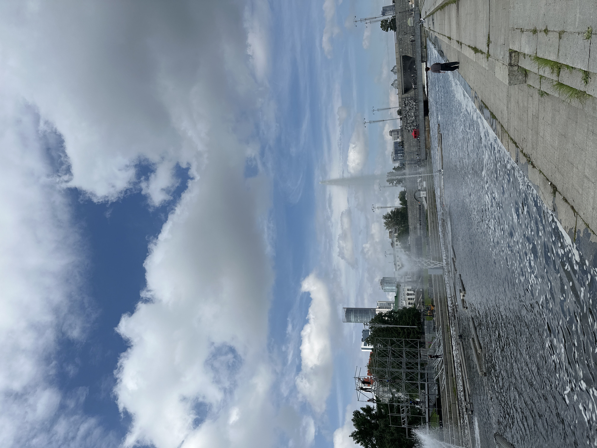 This is a photograph taken from the embankment of the Iset River in Yekaterinburg, Russia. The image shows a wide river with several fountains spraying water into the air. In the foreground, there is a concrete embankment with a single person standing and facing away from the camera. The river is dark and choppy, with some foam on the surface. Across the river, there are buildings, trees, and a large bridge. In the background, a tall, cylindrical skyscraper dominates the skyline. The sky is blue with large, white, fluffy clouds. There is scaffolding visible on the left side of the image, suggesting ongoing construction or maintenance work.