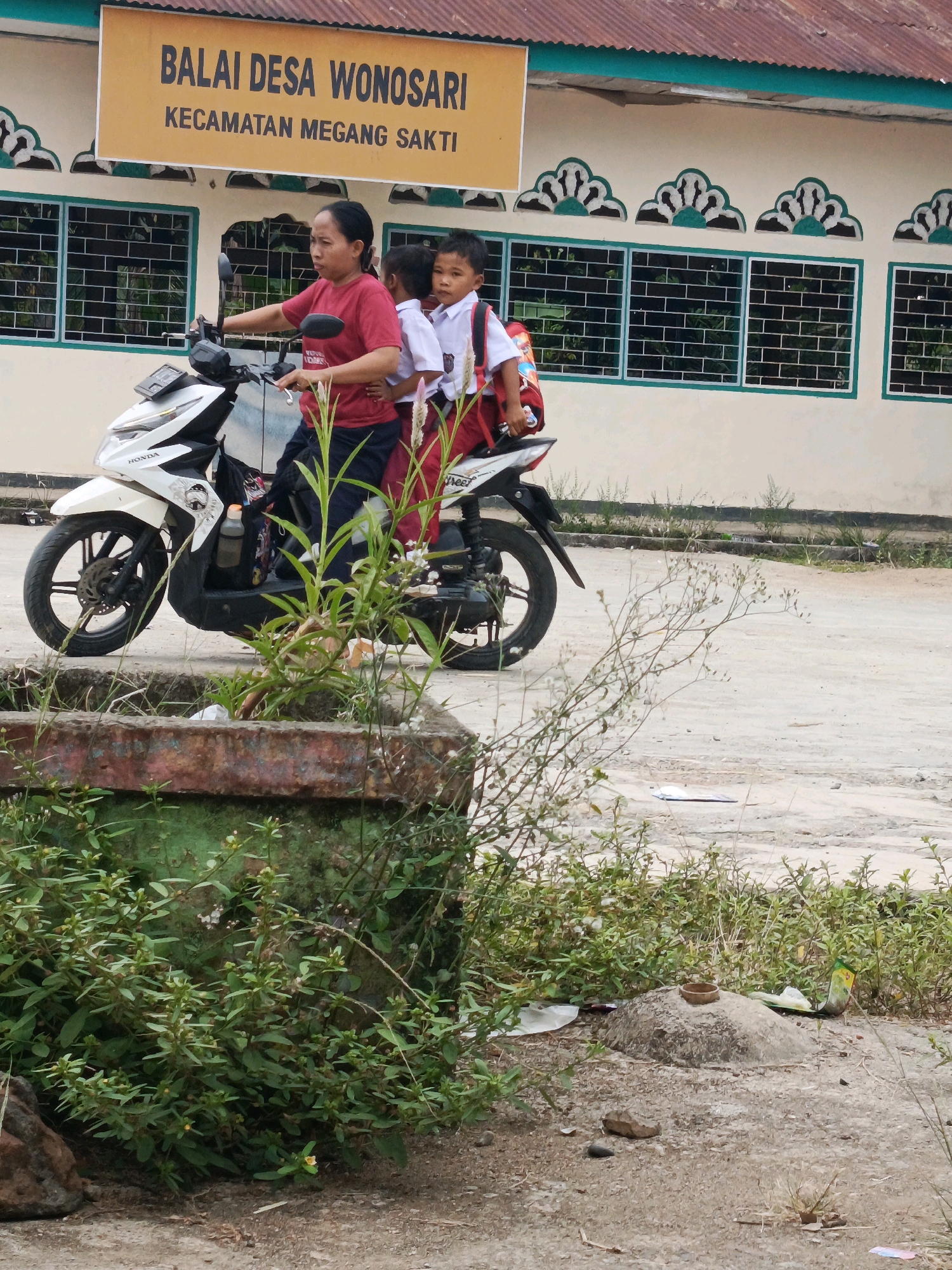 A woman in a red shirt is riding a white motorcycle in front of a building with a yellow sign that says "BALAI DESA WONOSARI KECAMATAN MEGANG SAKTI." She is carrying two young children, both wearing white shirts and backpacks. The children appear to be school-aged. The motorcycle is parked on a dirt or gravel surface, with some greenery and a concrete structure in the foreground. The building has windows with green trim and decorative elements above them. The sky is not visible. The scene appears to be outdoors in an Indonesian village.