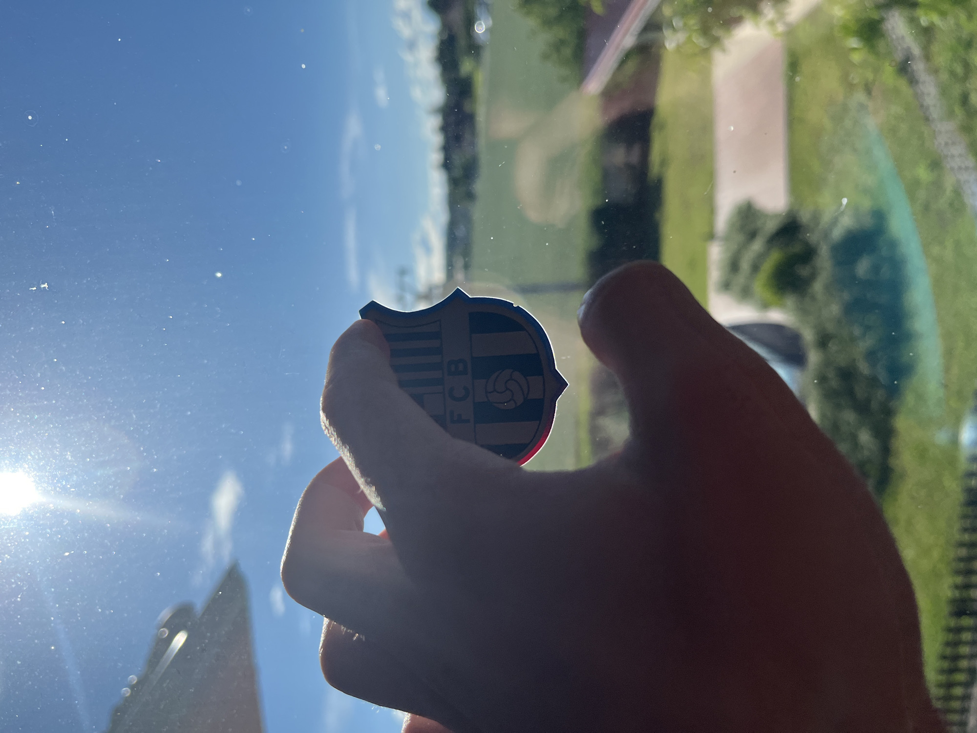 A hand is holding a blue and red shield-shaped emblem with the letters "FCB" and a soccer ball design against a bright blue sky with the sun shining overhead. The emblem is partially silhouetted by the sun. In the background, visible through a window, is a green field with trees and buildings in the distance.