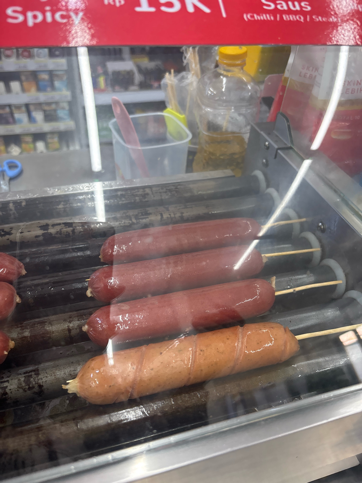 The image shows a close-up of a hot dog roller grill inside a convenience store in North Jakarta, Indonesia. Several hot dogs are on the grill, with some appearing cooked and others in various stages of preparation. In the background, shelves stocked with goods, including bottled drinks and packaged snacks, are visible. A clear plastic container with a pink utensil is also present. The overall scene suggests a food service area within a retail environment.