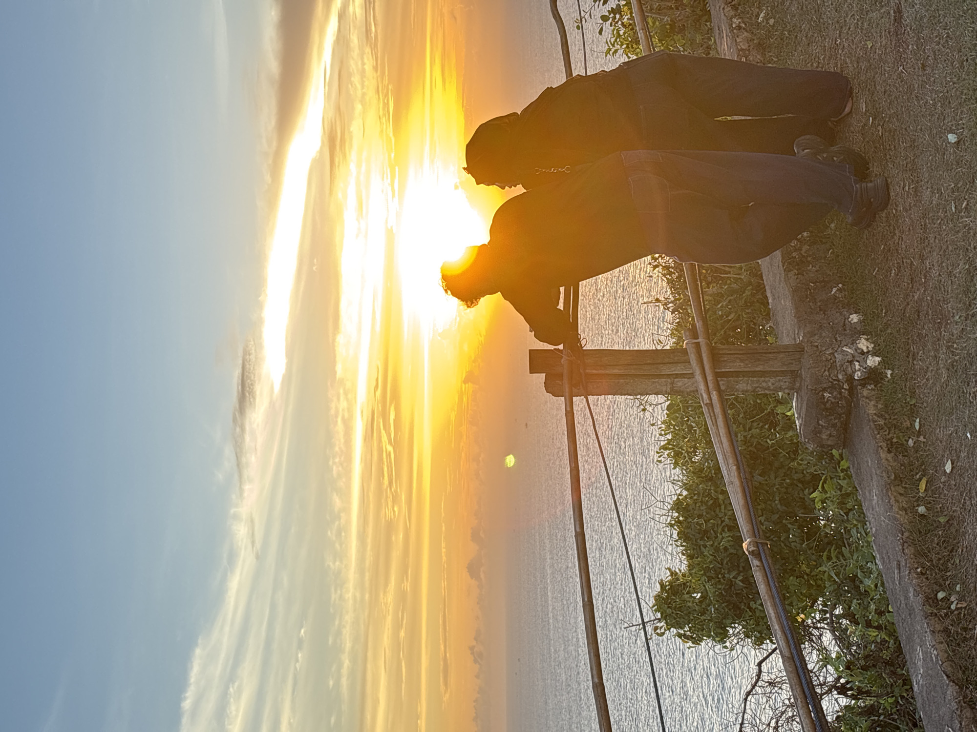 Two people are seen from the mid-torso up, standing on a grass-covered area, leaning over a wooden railing, and facing the sun setting over the ocean. The location is presumably in the city of Kuta Selatan, Indonesia. The foreground includes a section of the railing, some greenery, and a concrete ledge. The background comprises the ocean, sky, and setting sun, with bright, golden light. The two people are in silhouette.