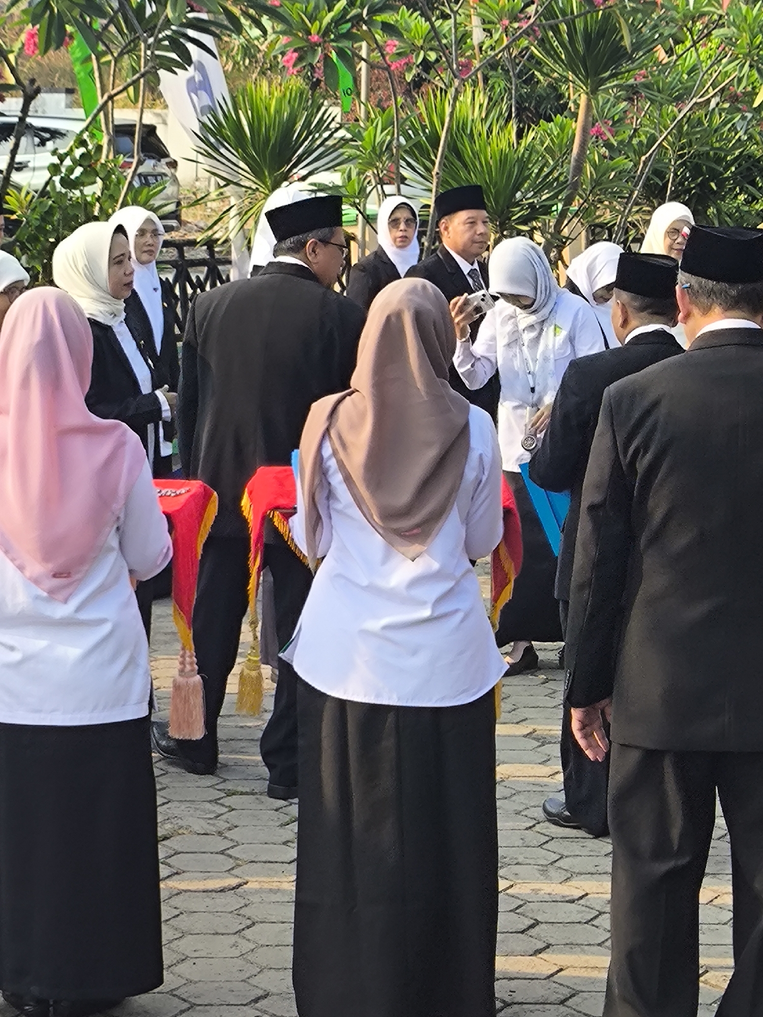 A group of people, predominantly women wearing hijabs and white shirts, and men in black suits and peci hats, are gathered outdoors. Some individuals are looking at their phones, while others appear to be observing or participating in an event. Behind the group, lush green trees with pink flowers are visible, suggesting a tropical or subtropical environment. The ground is paved with hexagonal tiles. Based on the provided context of the city of Cilegon, Indonesia, this scene likely depicts a formal gathering or ceremony taking place in a public space within the city.