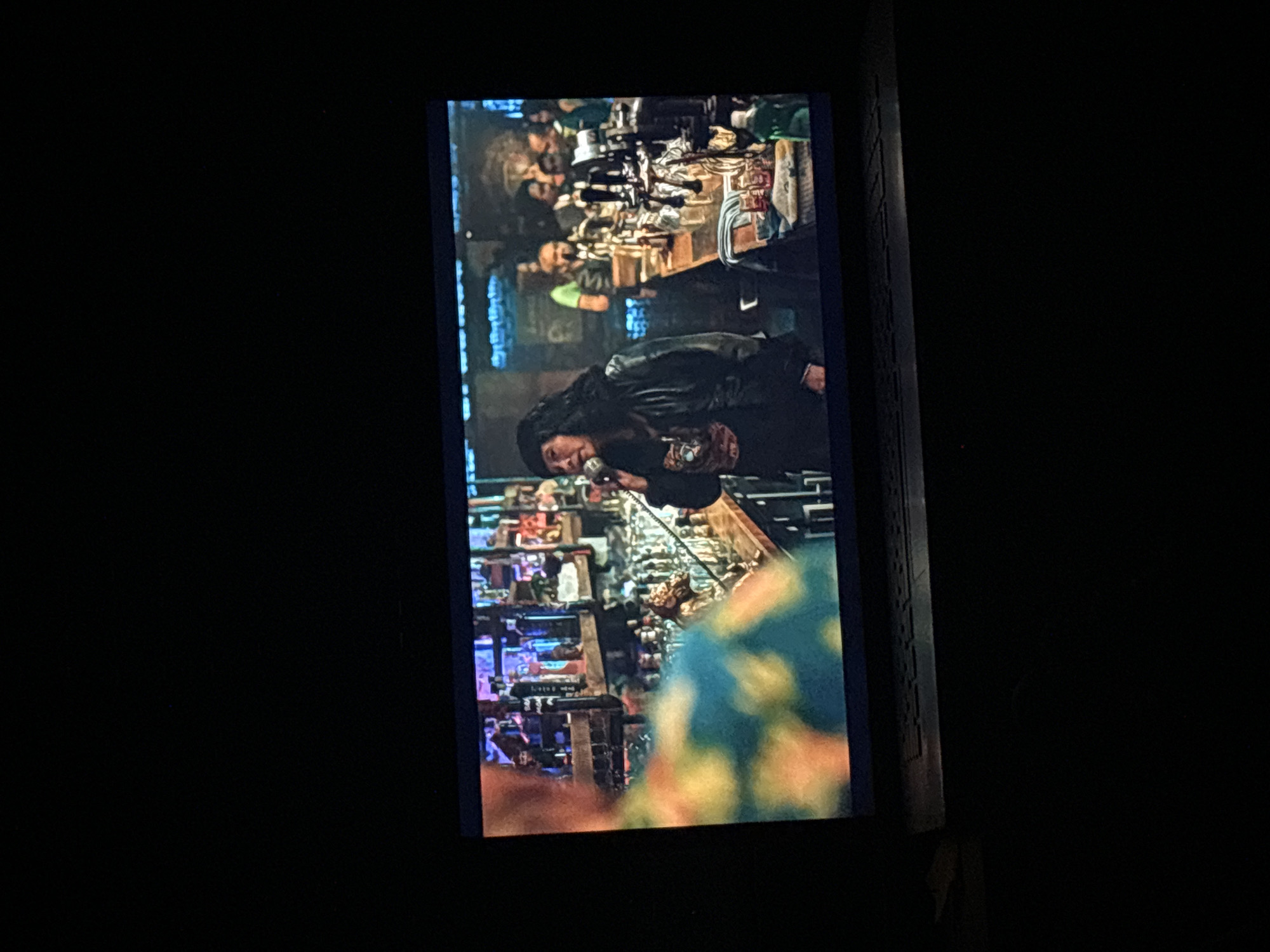 The image captures a vertically oriented screen displaying an indoor scene, possibly a bar, club, or performance venue. The primary subject is a person with long, dark hair, wearing a dark jacket or coat, holding a microphone and appearing to sing or speak into it. Their head is tilted slightly forward, suggesting an engaged performance.
Behind the performer, a bar or counter is visible, densely packed with numerous bottles, glasses, and various items characteristic of a drinking establishment. Several other individuals are present in the background, some seated or standing at the bar, observing the activity. Their features are indistinct due to the lighting and resolution.
The setting is dimly lit with artificial lights, implying a nighttime or late evening event. Colorful, blurred patterns appear in the lower foreground of the displayed scene, possibly a part of the decor or an object close to the camera. The overall atmosphere suggests a social gathering centered around a performance. No discernible text is visible within the image. The image lacks specific visual identifiers for Budva, Montenegro.