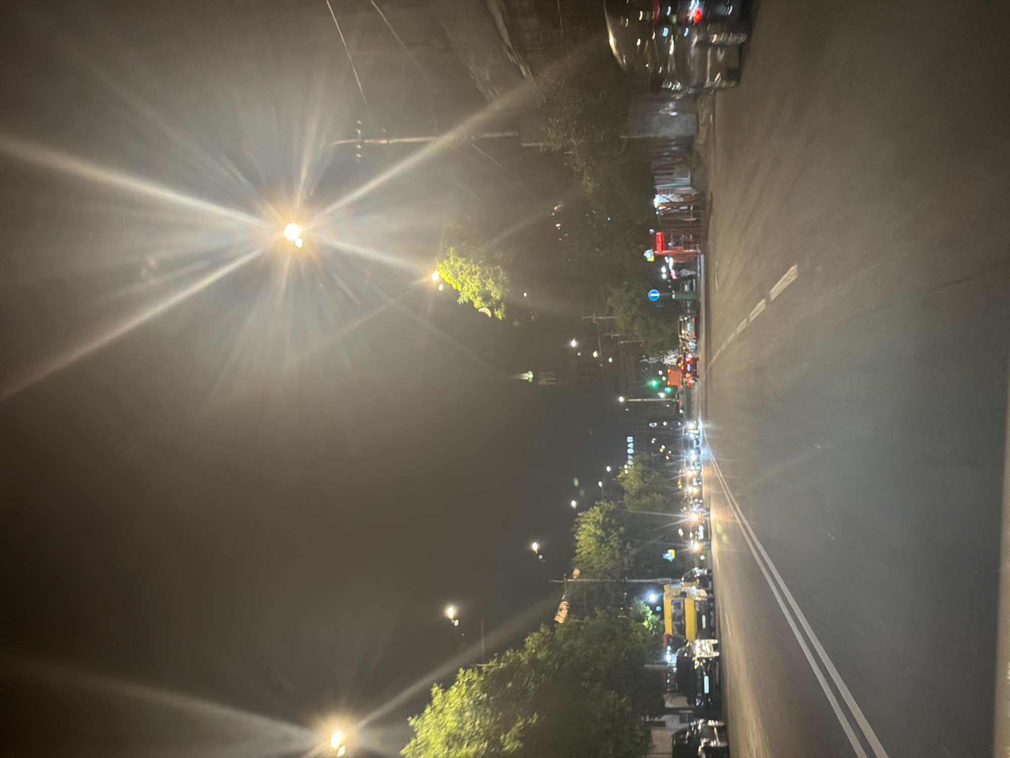 This image depicts a nighttime street scene in Yerevan, Armenia, likely on a busy avenue. The perspective is from a vehicle moving forward, evidenced by the road lines and the foreground. Several vehicles are present on the road, including a yellow van, black cars, and a bus, some of which have their headlights on. Streetlights and building lights illuminate the scene, creating lens flares and a hazy atmosphere, particularly from the bright streetlights overhead. Trees with green foliage line the sides of the road, and in the distance, a building with a spire is visible. The presence of parked cars on the left side of the road and the general urban setting suggest a populated area within the city. The overall impression is of a typical urban night drive.