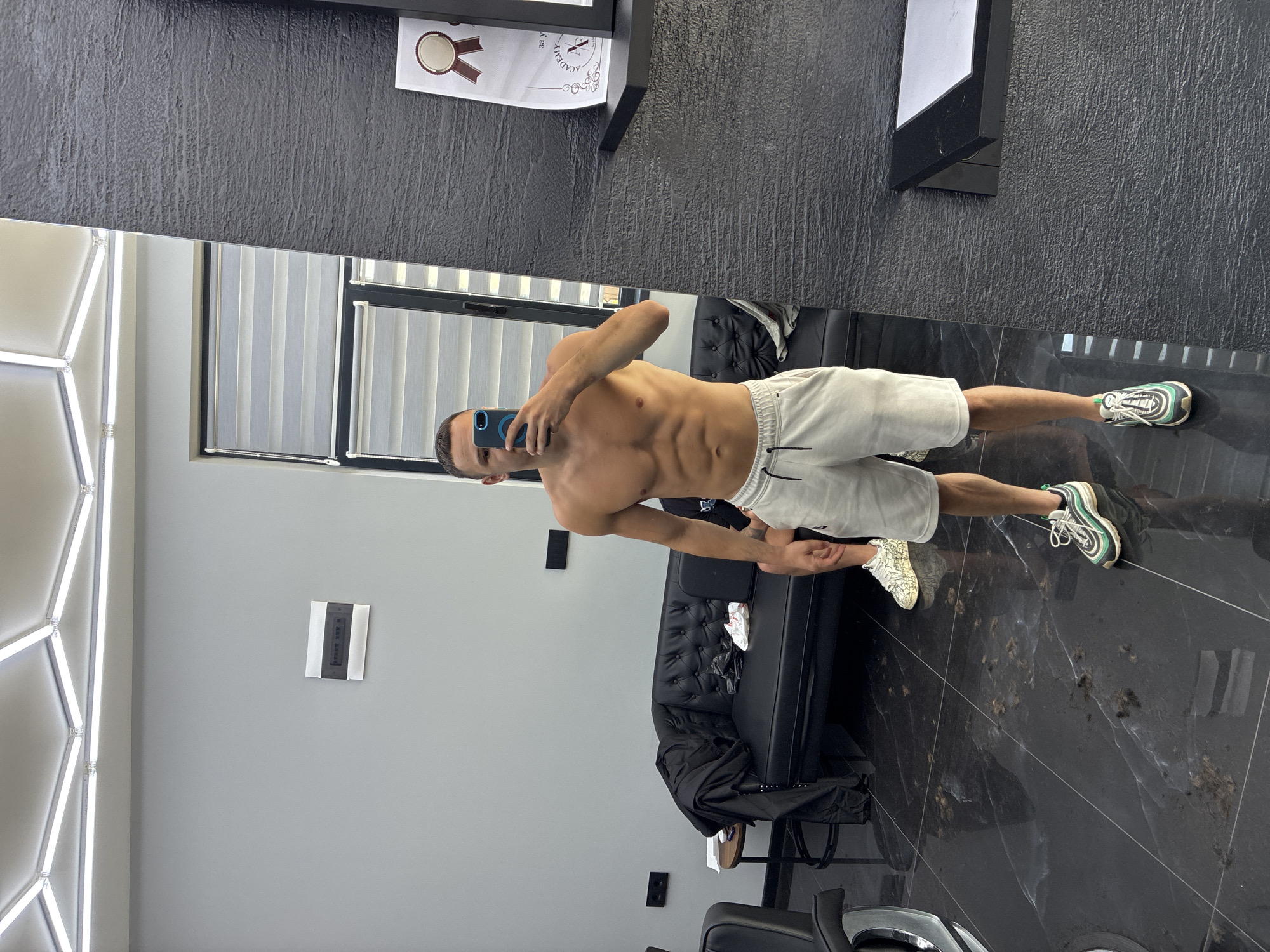 A man is depicted taking a mirror selfie in an indoor setting, likely in Simitli, Bulgaria. He stands shirtless, showcasing an athletic build, and wears light-colored shorts with a white drawstring. On his right foot, he wears a prominent green, black, and white sneaker, while his left foot sports a white and beige sneaker with a textured pattern. He holds a dark blue smartphone up to capture his reflection.
The room has a modern and sleek aesthetic. The floor is covered with dark, polished tiles or marble, featuring subtle white and gold veining. The walls are a mix of light grey and a textured, dark grey or black material. A distinctive, geometric light fixture with bright white LED strips is mounted on the light grey wall.
In the reflection, a large window with vertical blinds is visible, suggesting daylight illumination. Other reflections show a black tufted sofa or bench and what appears to be another black chair, reinforcing the professional or upscale casual environment.
On the actual wall above the mirror, a piece of paper is taped, displaying a certificate with a seal design and the partially visible Cyrillic word "АКАДЕМИЯ" (ACADEMY), indicating a professional or training academy context. Another small, dark rectangular object is mounted on the grey wall, but its text is illegible. The overall scene suggests a personal moment captured in a stylish, possibly a salon, gym, or studio space.