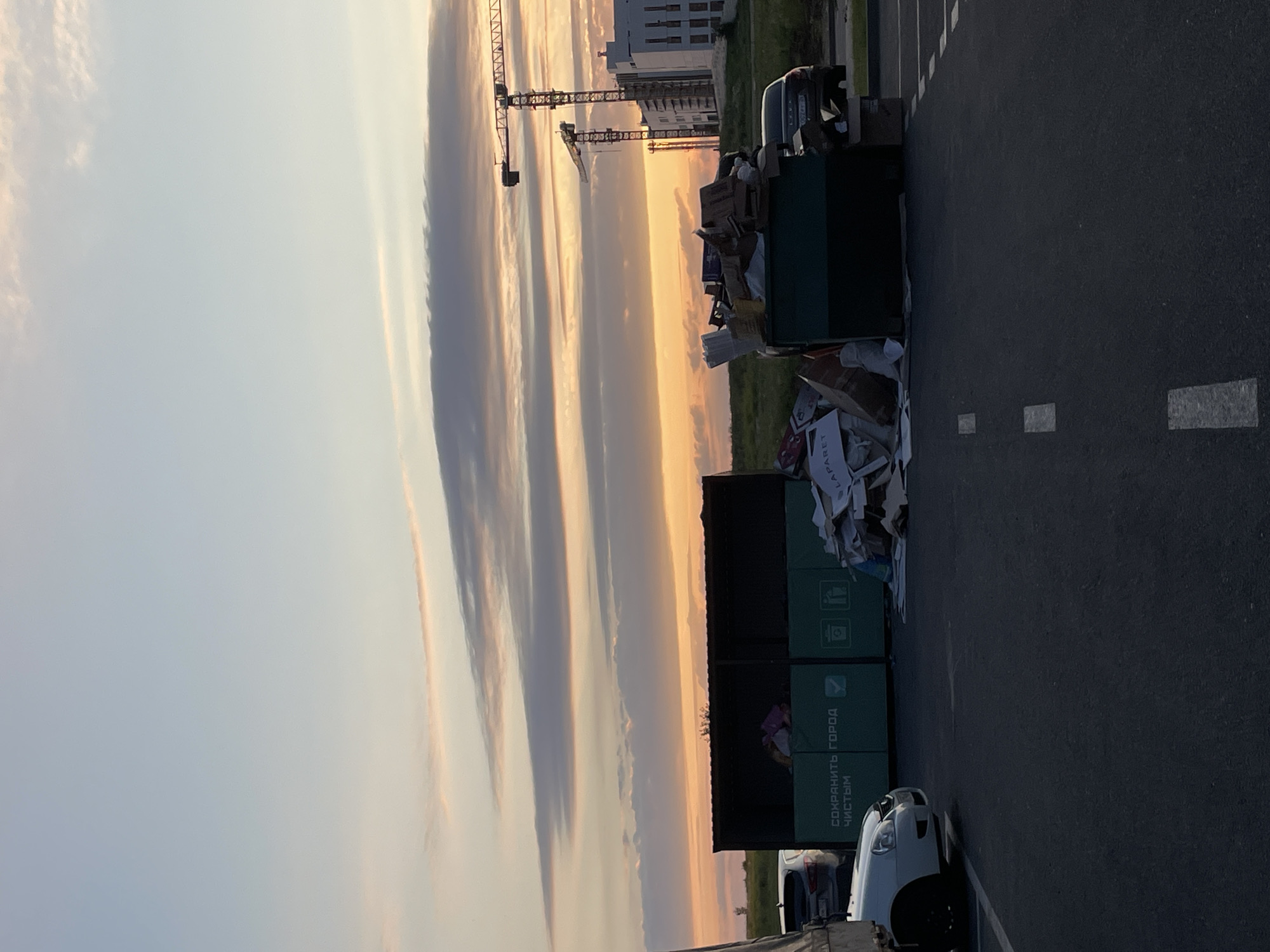 The image depicts an outdoor scene at dusk, likely in the city of Stavropol, Russia. In the foreground, a section of asphalt road with white lane markings is visible. To the left, a white car is parked. Further back, two large green dumpsters are situated. One dumpster has text in Russian that translates to "Keep the city clean." It also features icons for recycling and waste disposal. This dumpster is partially filled with trash bags. Adjacent to it is another dumpster overflowing with cardboard boxes and other debris. To the right of the dumpsters, a dark-colored car is parked. In the background, construction cranes and modern white buildings are visible against a sky filled with wispy clouds illuminated by the setting sun, which casts an orange glow. The overall scene suggests a construction site or a residential area undergoing development, with a focus on waste management. There are no people or animals visible in the image.