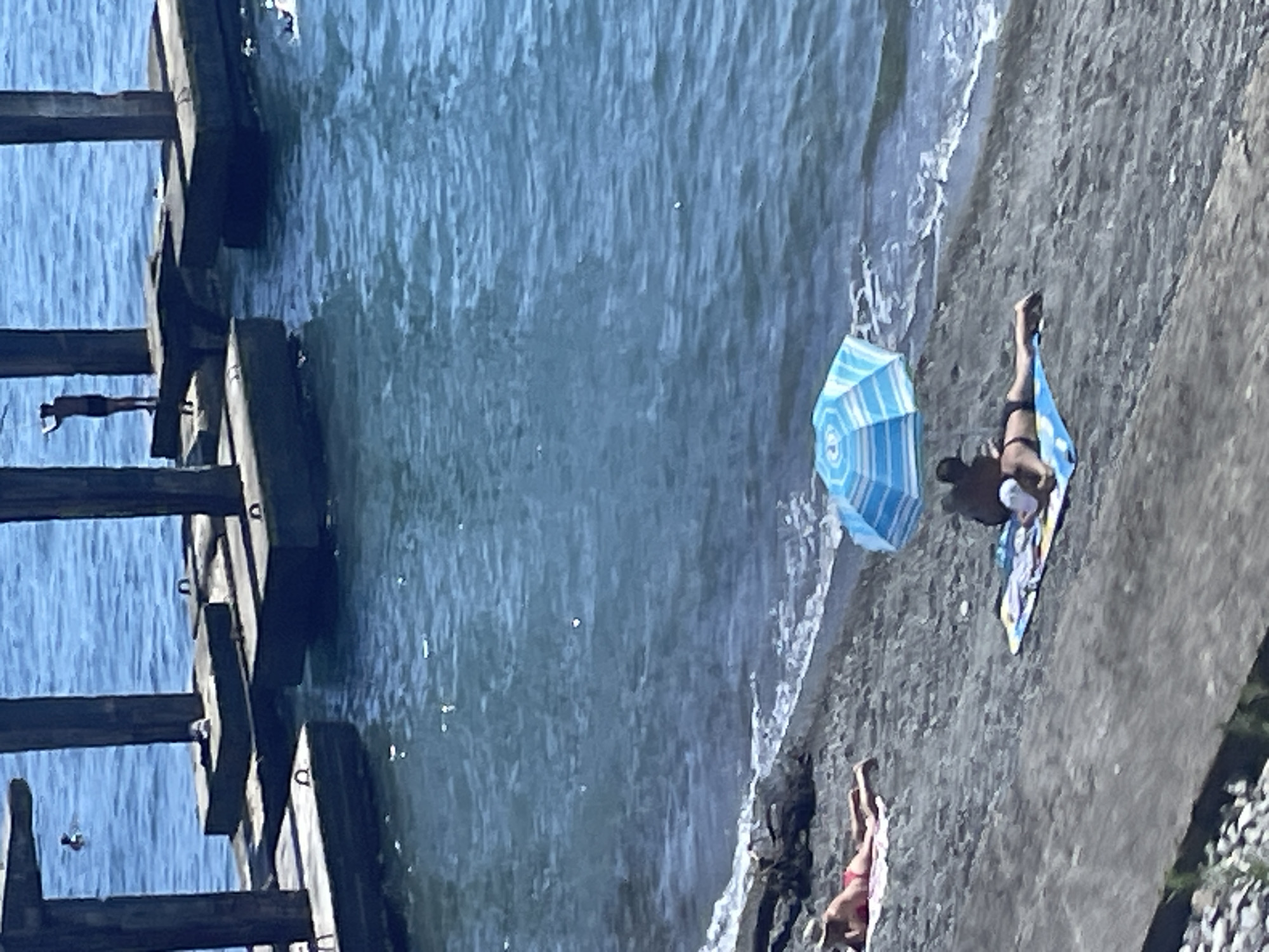 The image depicts a sunny day at a beach with a pier structure in the background and people sunbathing on the shore. The pier, constructed with concrete pillars and platforms, extends into the calm blue water. Several individuals are visible on and around the pier, with one person standing on a platform, appearing to be observing the water or preparing to jump. Another person is swimming in the distance.
On the dark pebble beach in the foreground, two individuals are sunbathing. One person is lying on a towel under a blue and white striped umbrella, facing away from the camera. Another person is lying on a towel nearby, also facing away from the camera.
The water is clear and blue, with small waves gently lapping at the shore. The beach is composed of dark pebbles and sand. The overall scene suggests a relaxed summer day at the coast. Given the architectural style of the pier and the general appearance of the coastline, this location is likely a public beach in the city of Sochi, Russia, known for its numerous piers and seaside resorts.