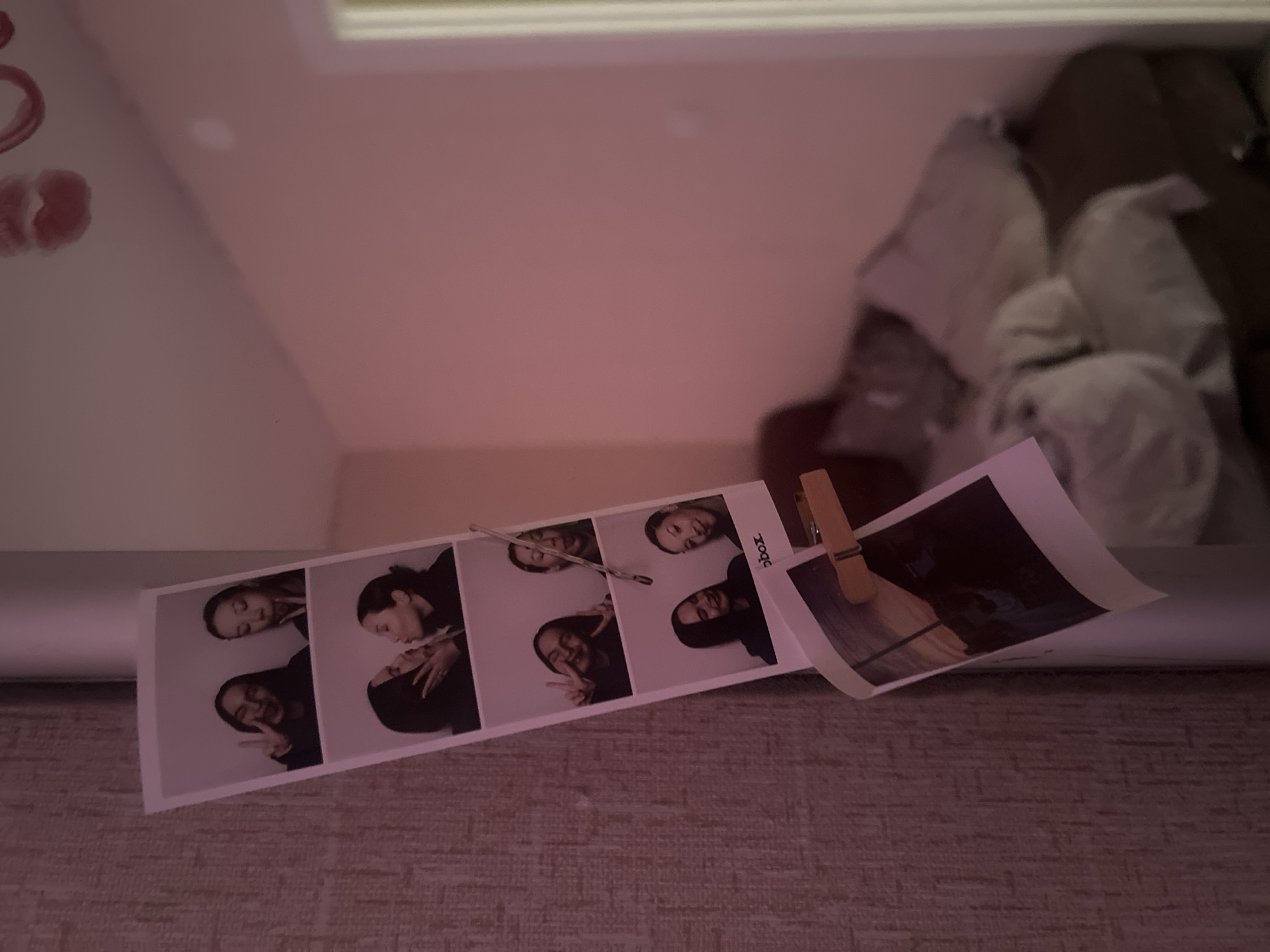 The image shows a photo strip held by a clothespin on a surface with a textured, possibly carpeted, covering. The photo strip contains four images of two individuals, each with different poses and expressions. To the right of the photo strip is another photograph, held by the clothespin, featuring a less distinct subject in dark tones. The background consists of a light pink wall and what appears to be a pile of soft objects or bedding, potentially a pillow or comforter, situated in the upper right. Based on the details provided in the prompt, the location is Chaikovsky, Russia.