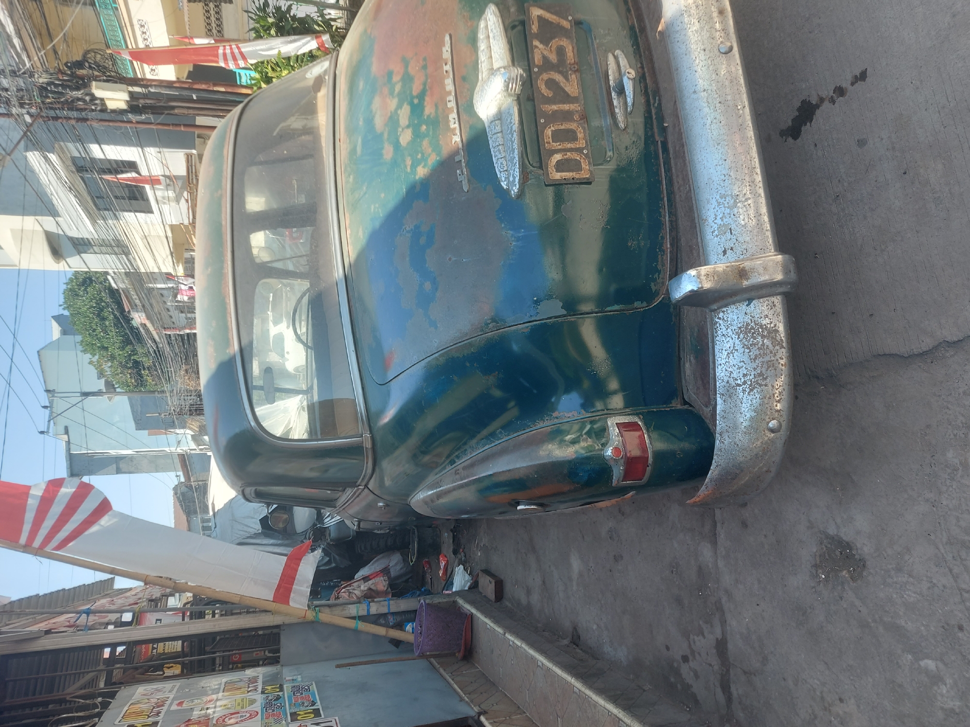 An aged, dark teal Plymouth car, possibly a 1950s model, is parked outdoors on a concrete surface. The car exhibits significant rust and paint degradation, particularly on the trunk and rear bumper. A license plate with the characters "DD1237" is visible. To the left of the car, a narrow walkway runs alongside a building adorned with various stickers and posters. A bamboo pole supports a long, white flag with red stripes. In the background, buildings and utility poles suggest an urban setting, with glimpses of other flags and vehicles. The overall impression is of a street scene in a tropical climate, potentially in Makassar, Indonesia, given the context.