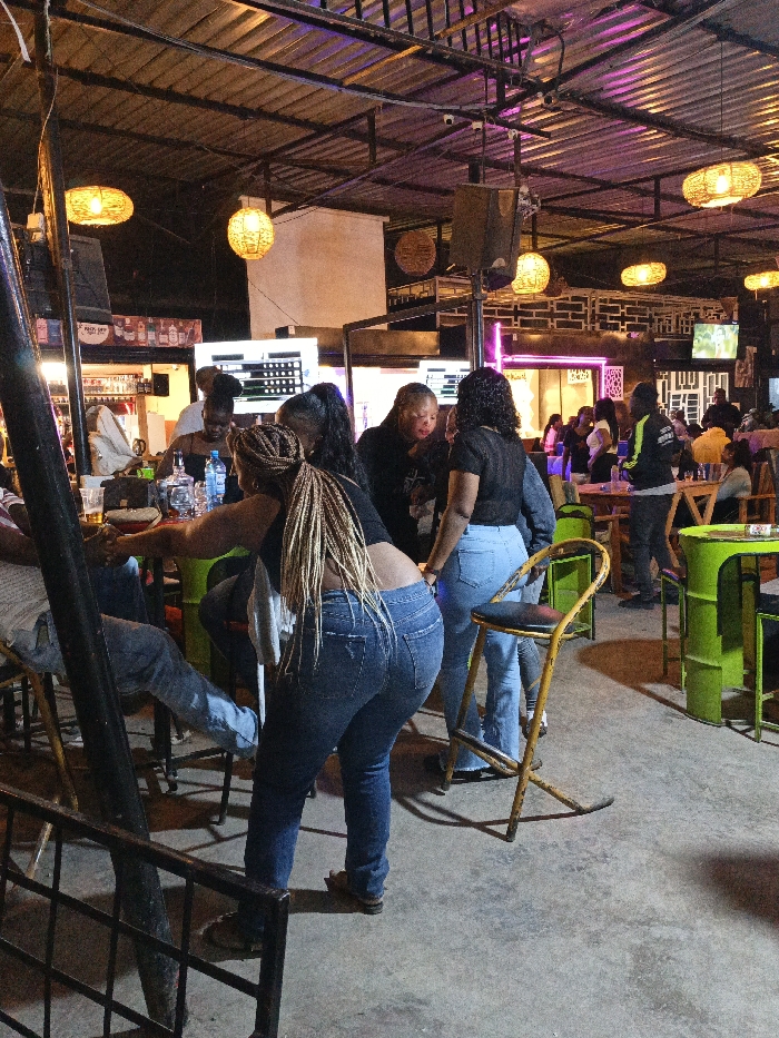 At an outdoor venue, a group gathers around a bar. The bar is illuminated by screens and overhead pendant lights. Several women are visible, wearing casual clothing like jeans and dark tops. Some are standing, while others are seated at high stools. Further back, other patrons sit at tables, and a television screen glows in the background. The flooring is concrete, and the overall ambiance is relaxed and convivial.