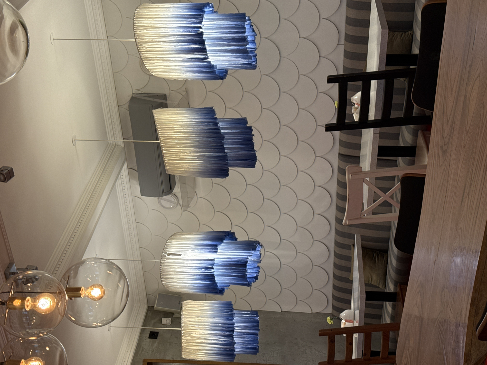 The image displays the interior of a stylish commercial establishment, likely a restaurant or cafe, characterized by its distinctive lighting and decorative elements. The scene is unoccupied, presenting a calm and composed atmosphere.
From the white ceiling, adorned with classical crown molding, hang several unique light fixtures. Two large, clear spherical glass pendant lights with visible incandescent bulbs emit a warm glow. In contrast, four tiered pendant lights, possibly made of pleated fabric or paper, showcase an elegant ombre effect, transitioning from white at the top to a deep blue at the bottom. These shades cast a cool, diffused light. A grey wall-mounted unit, possibly an air conditioner or speaker, is visible near the ceiling.
The left wall is a focal point, featuring a textured design composed of overlapping white semi-circular tiles, creating a striking fish-scale or scallop pattern.
The furniture includes booth seating upholstered in gray and white stripes, providing a comfortable option. Various chairs are visible, including a black ladder-back chair and a white cross-back chair, complementing the mixed aesthetic. A light wooden table or counter runs along the bottom right. On one of the tables, a small white figurine holding a yellow flower adds a delicate decorative touch.
The overall impression is one of modern design with a refined and inviting ambiance, utilizing distinct lighting and wall textures to create a unique space in Perm, Russia. The illumination from the pendant lights suggests an indoor setting where artificial lighting is primary. No visible text is present.