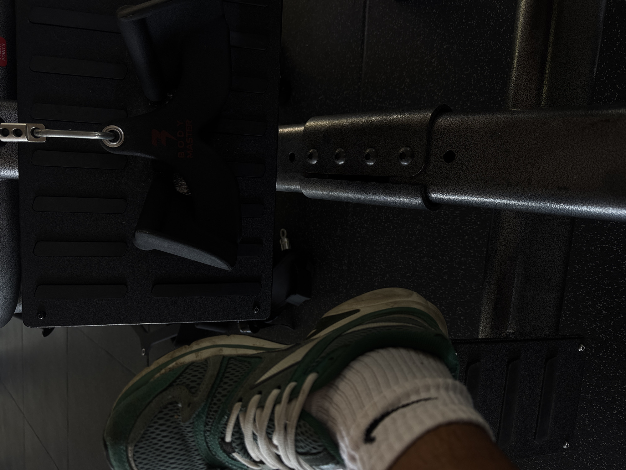 A close-up, low-angle shot shows a person's foot in a green and white athletic shoe and white sock stepping onto the footplate of a black weight machine. The machine has a black textured surface with two black rubberized grips attached to a metal cable. The brand "BODY MASTER" is visible on the grips. The scene is indoors, on a dark, speckled floor, typical of a gym environment. The image is underexposed, with the focus on the shoe and the machine. The location is Kuta Selatan, Indonesia.