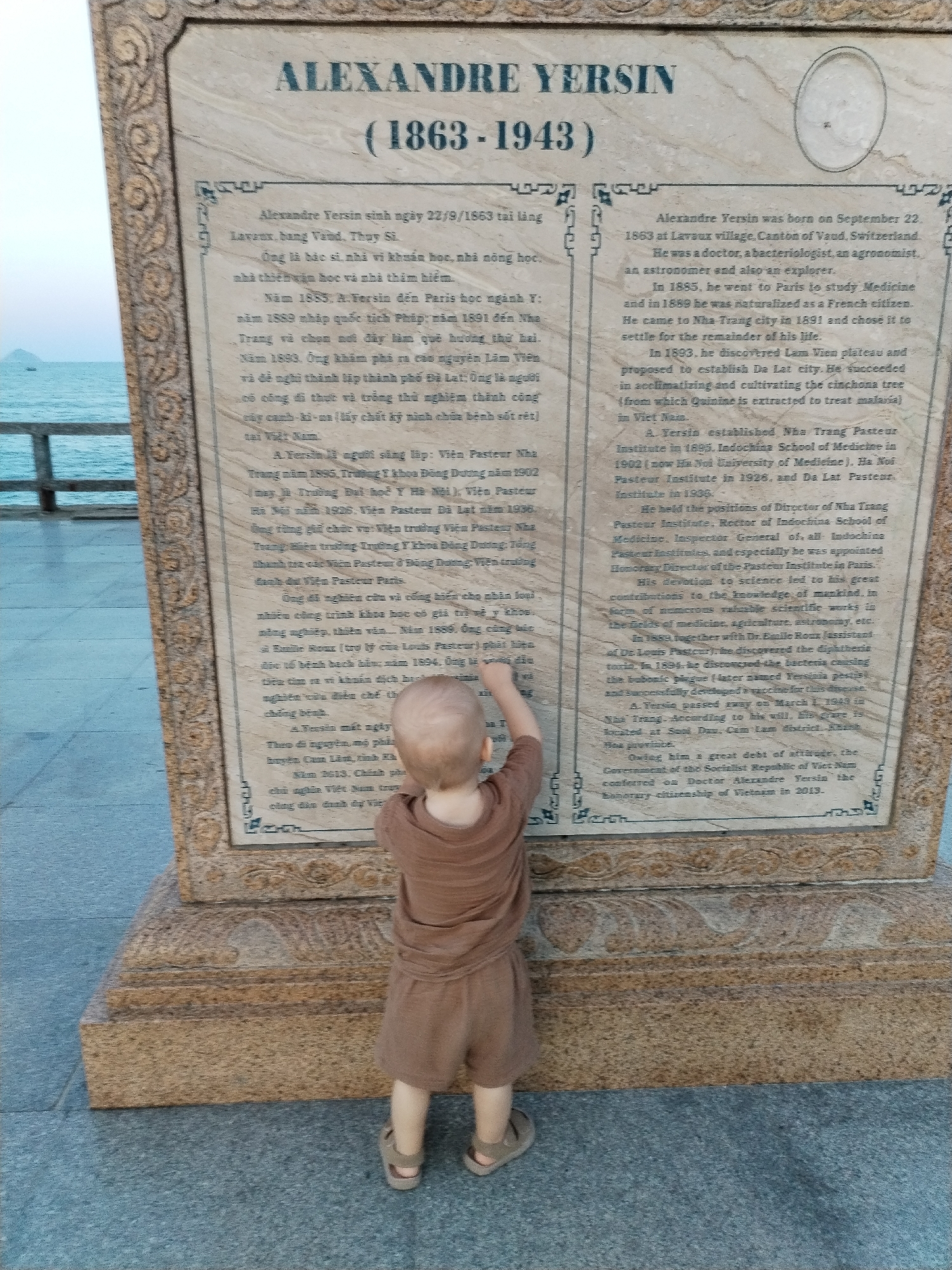 A full shot shows a light-skinned toddler in brown clothing looking up at an upright, waist-high, tan plaque. The plaque has inscriptions on it written in two languages, Vietnamese and English, in black writing. Text at the top of the plaque reads "Alexandre Yersin" followed by "(1863-1943)" underneath. The plaque rests on a light brown pedestal. In the background behind the plaque and the toddler is a view of the ocean and a cloudy sky. The setting is Nha Trang Ward in Vietnam.