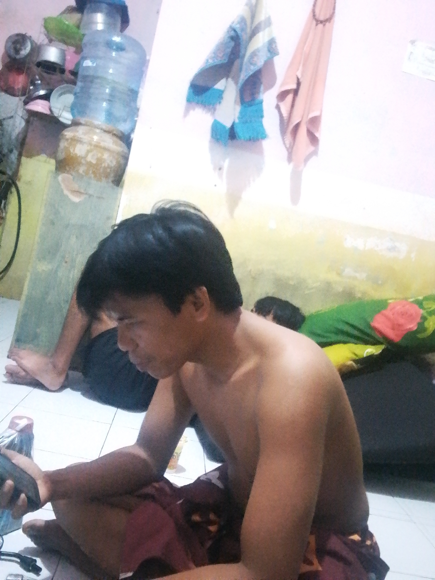 This media file features a young man sitting on a tiled floor, facing left. He is shirtless and wearing maroon shorts. He is holding a dark object, possibly a phone or gaming device, in his hands and appears to be looking at it. His expression is neutral, and the lighting suggests it might be daytime or an indoor setting with artificial light.
In the background, to the left, is a stack of water bottles and several containers, including what appears to be a large clay pot and metal bowls. A green wall is visible behind the man, and to the right, there is a person lying down on a green and yellow patterned mattress with a floral-designed pillow. Hanging on the wall in the background are two pieces of fabric, one patterned with blue and white stripes and the other a solid peach color.
The setting appears to be a simple indoor living space, possibly a bedroom or common area, in Bohar, Indonesia. The overall atmosphere is casual and domestic, with no specific event or strong emotions being conveyed. There is no visible text in the image.