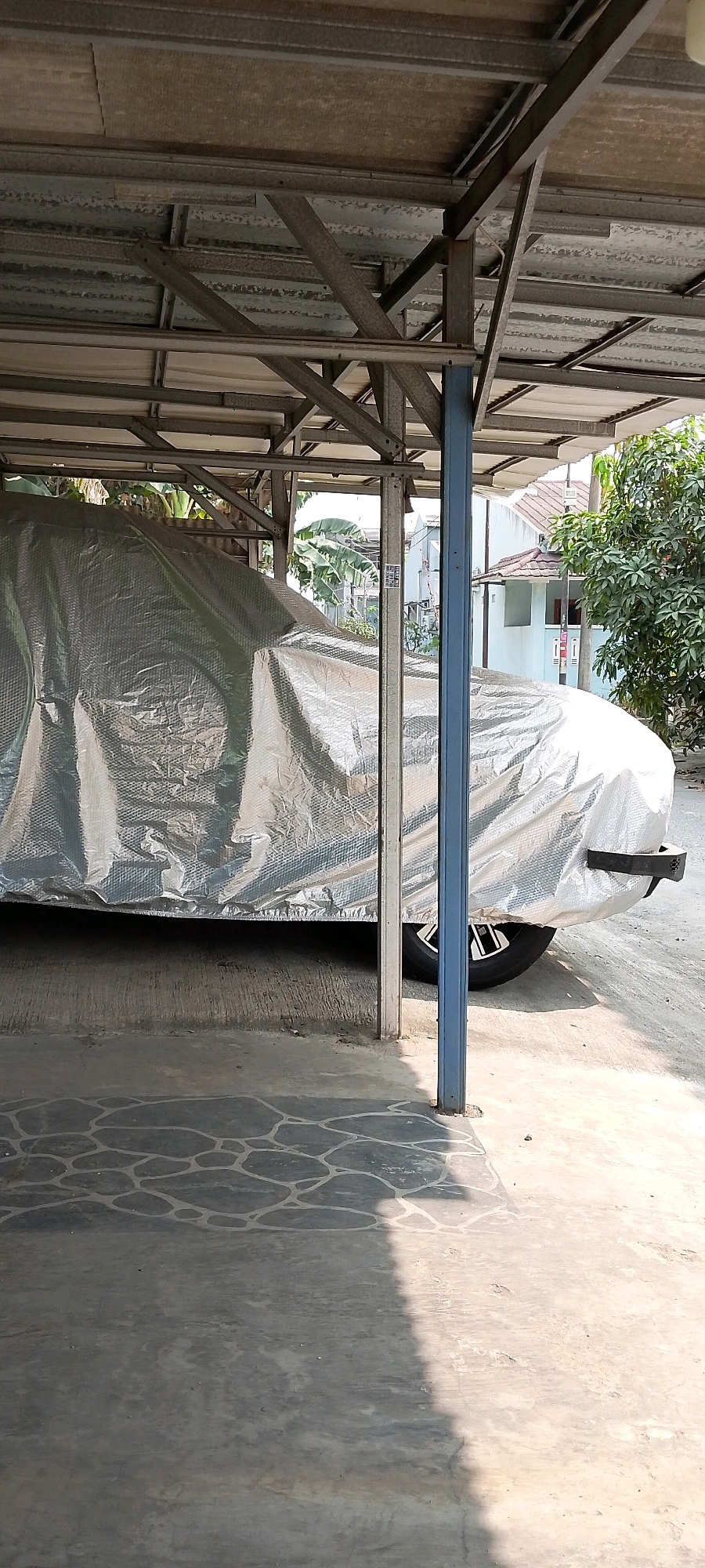 The image depicts a car covered with a silver-colored bubble wrap-style car cover, parked under a metal-framed open-air shelter. The shelter has a corrugated metal roof supported by a network of steel beams and a vertical blue metal pole. The ground beneath the shelter is concrete, with a section of decorative patterned paving in the foreground. Beyond the shelter, in the background, residential buildings with tiled roofs and trees are visible, suggesting an outdoor urban or suburban environment. The car's tire is partially visible, indicating it is a vehicle. The lighting suggests it is daytime, with shadows cast on the ground. No people are present in the frame. The overall scene is static and depicts a parked, covered vehicle in an outdoor parking area. The location is identified as Satriamekar, Indonesia.