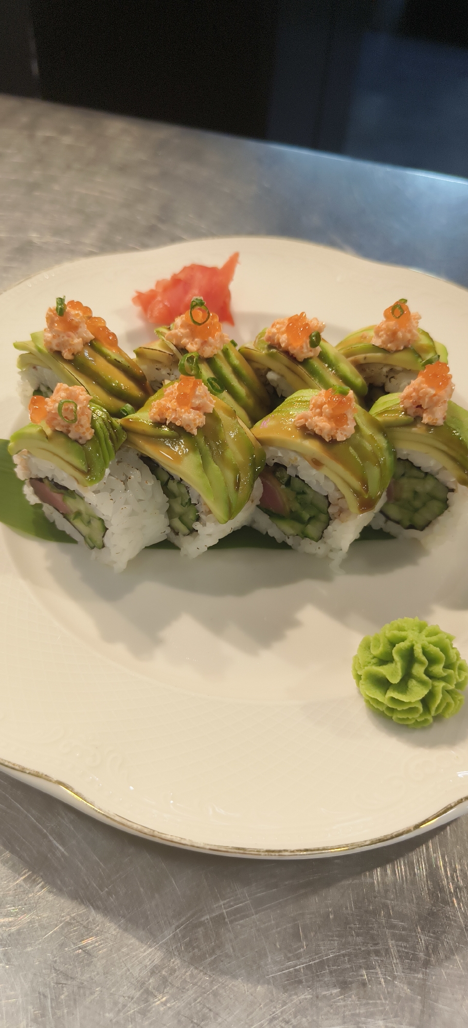The image shows a plate of sushi rolls, likely indoors in the city of Unknown, Unknown. There are eight sushi rolls arranged in a row on a white plate with a gold rim. Each roll is topped with sliced avocado, a pink topping, and a few green chives. Next to the sushi is a dollop of green wasabi and a portion of pink pickled ginger. The plate sits on a metallic surface.