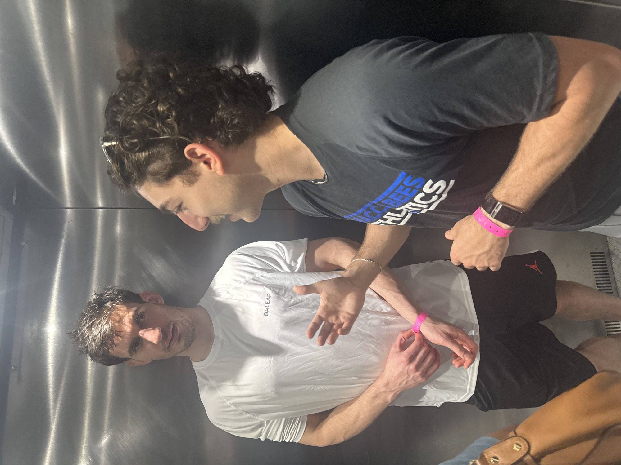 The image depicts two males in what appears to be an elevator. The elevator's interior has stainless steel walls. The man on the left wears a white t-shirt with the word "BALEAF" on it and black athletic shorts with a red Jordan logo. He is making a gesture with his right hand while looking up at the other man. He wears a pink wristband on his left wrist. The other man wears a dark t-shirt with light-colored lettering that includes the word "Athletics," and black athletic shorts. A watch with a black band and a pink wristband is visible on his left wrist. The specific location in Aventura, United States of America, cannot be determined from the image.