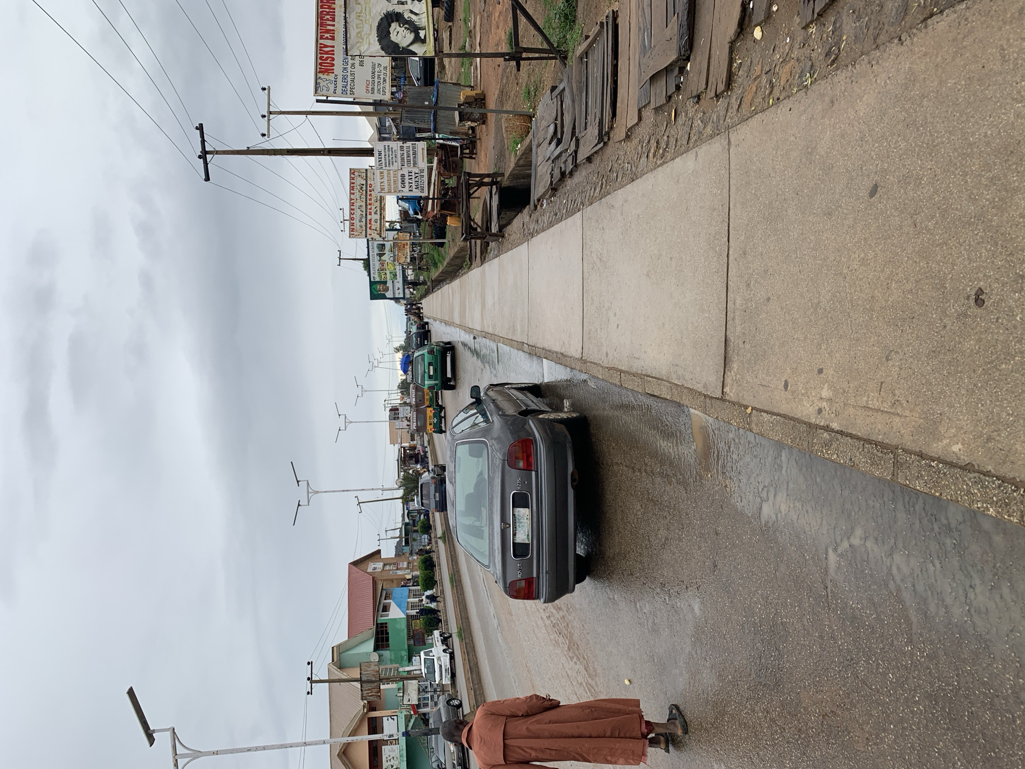 The image captures a street scene in Rafin Par, Nigeria. A gray Rover 623Si is parked on the right side of a wet asphalt road. A man wearing a brown robe is walking near the parked car. A concrete sidewalk runs parallel to the road, bordered by a drainage ditch covered with wooden planks. Utility poles with overhead power lines stretch along the left side of the road. Various billboards advertising local businesses, including "NOSKY ENTERPR" and "INNOCENT EMEKA," are visible in the background. There is moderate traffic on the road, consisting of cars and trucks. Buildings and trees line the left side of the street. The sky is overcast and gray.